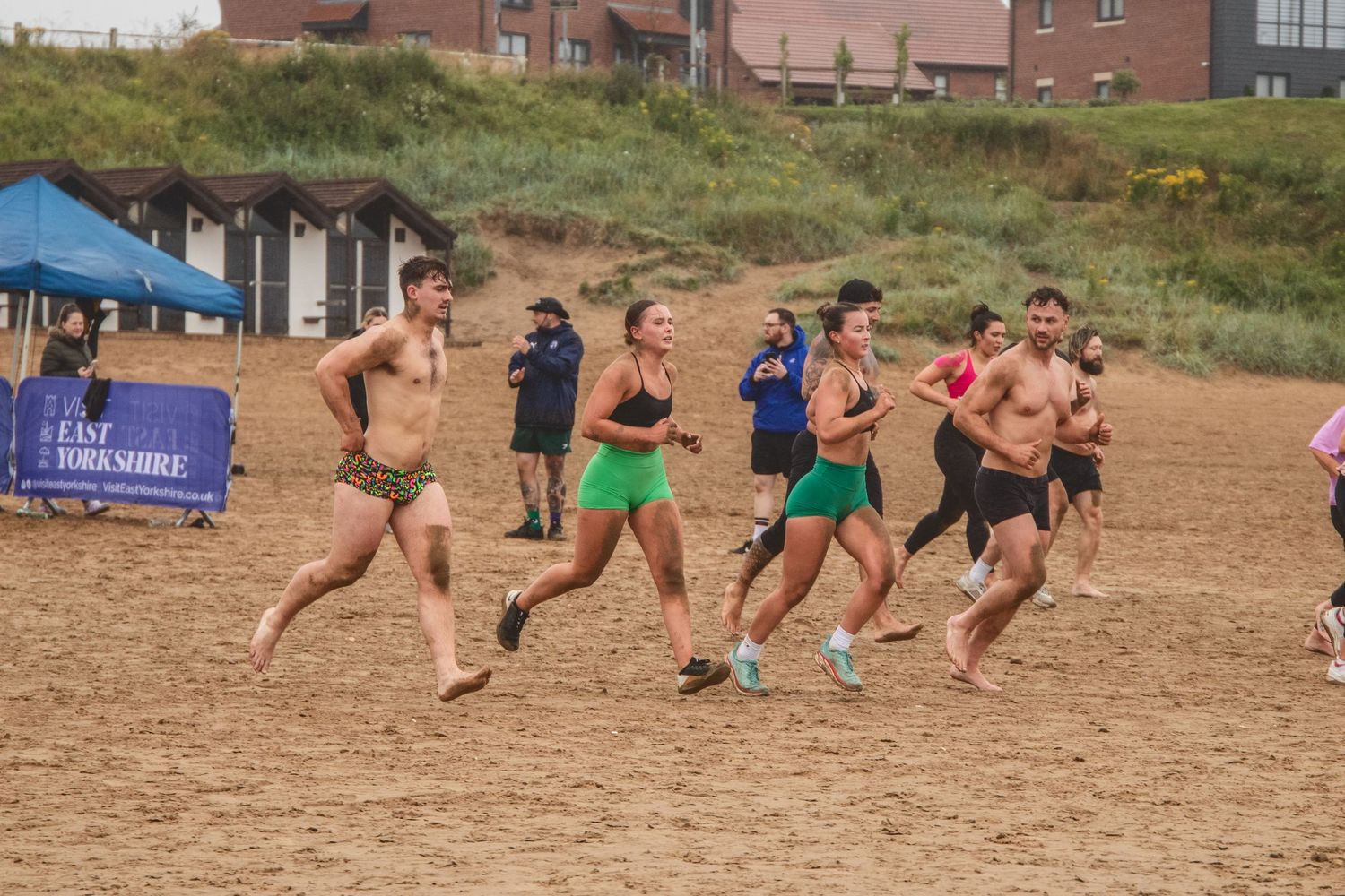 Athletes compete in a seaside running race on sandy beach terrain with spectators watching under blue tents in background.