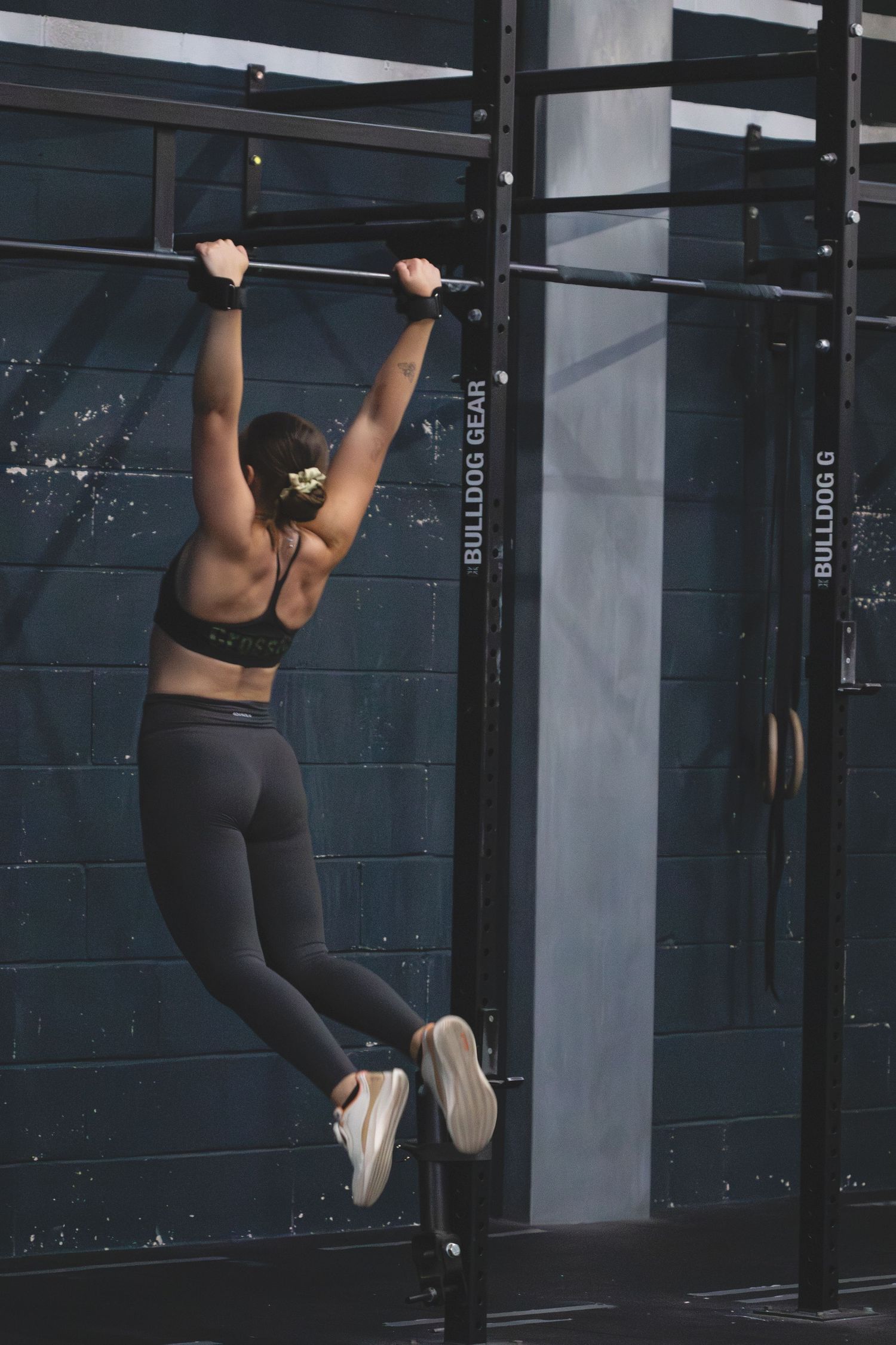Athletic person performs pull-ups on a horizontal bar in a dimly lit gym while wearing dark activewear gear.