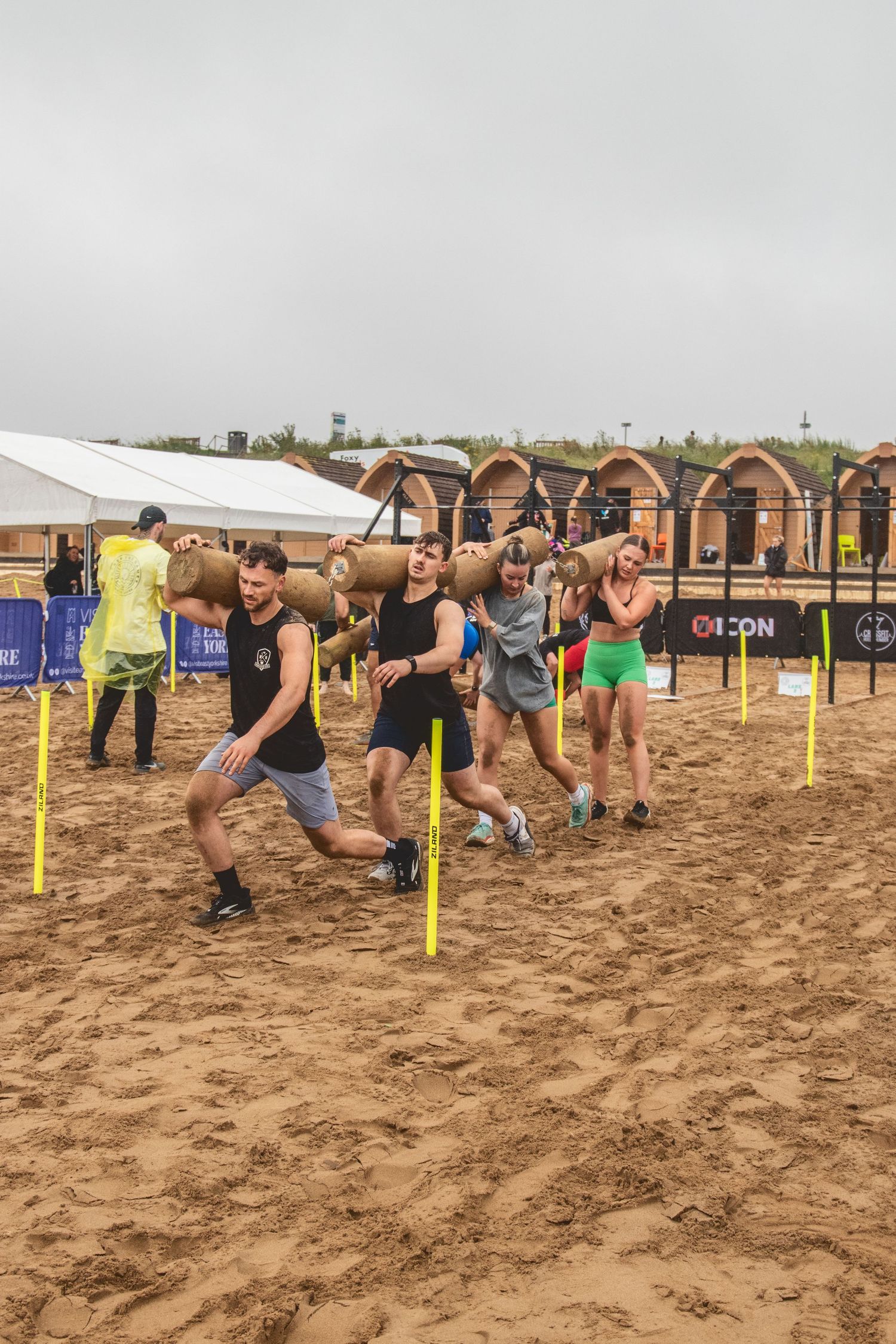 A group of runners participate in a muddy obstacle course race, crawling under barriers on a sandy terrain.