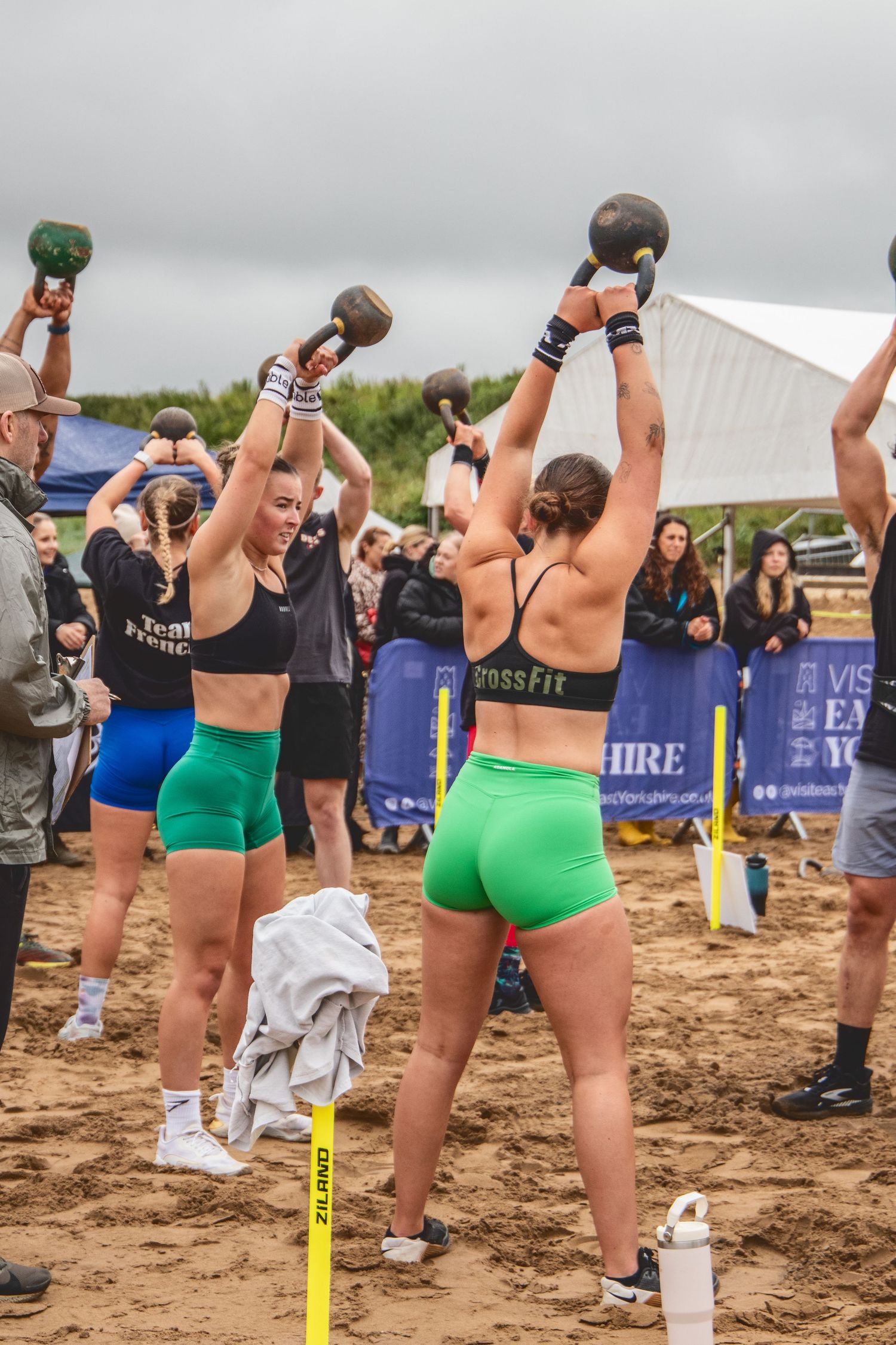 A group of athletes performing kettlebell exercises together on a sandy beach during an outdoor fitness competition.
