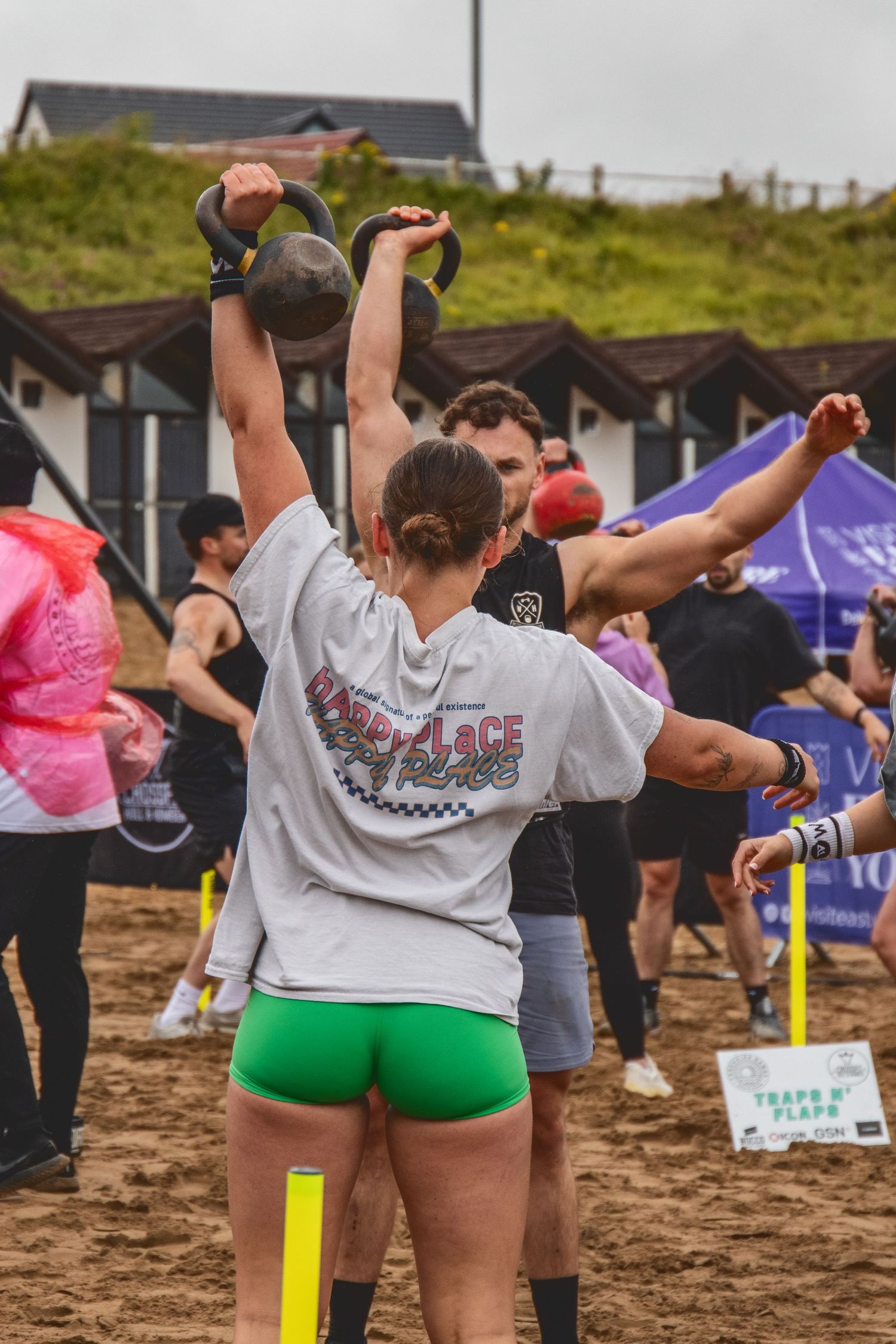 Athletes compete in an outdoor kettlebell competition event on sandy ground with tents and hills in the background.