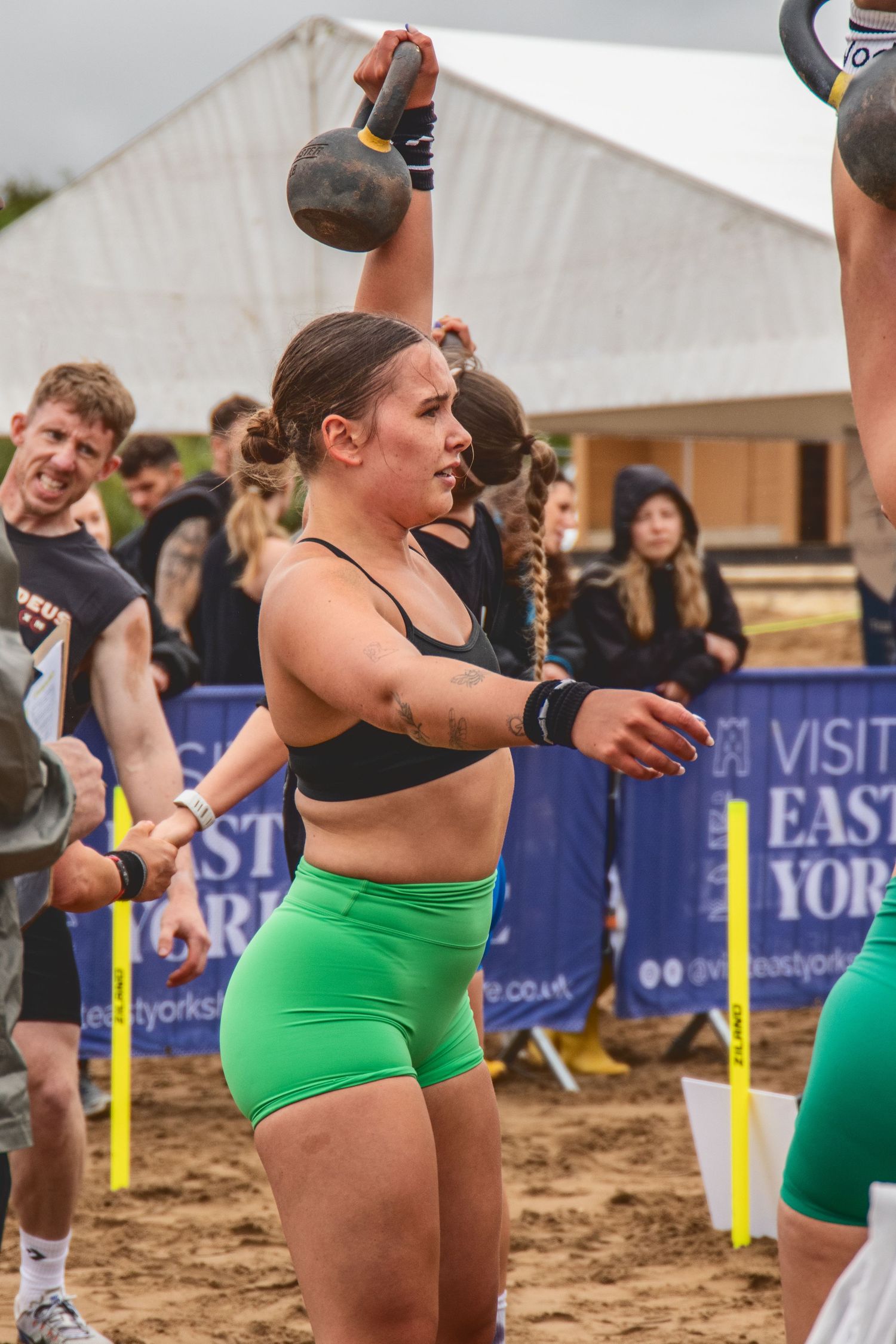 Athlete in bright green shorts performs kettlebell workout during outdoor beach competition.