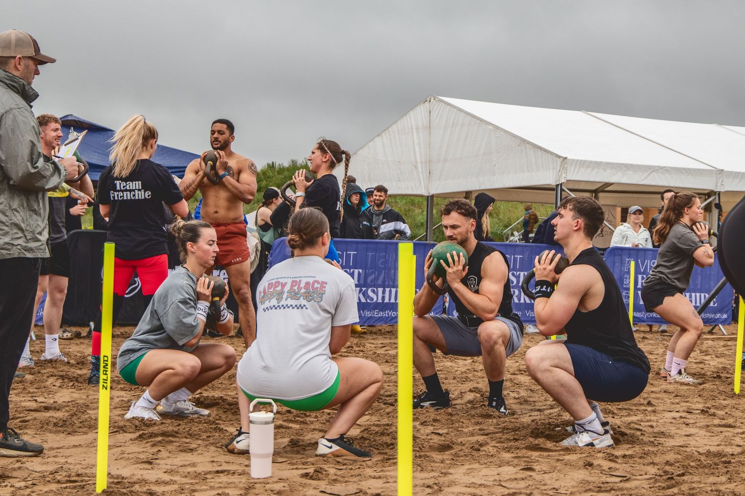 Athletes perform synchronized squats during an outdoor fitness competition on a sandy surface near a white tent.
