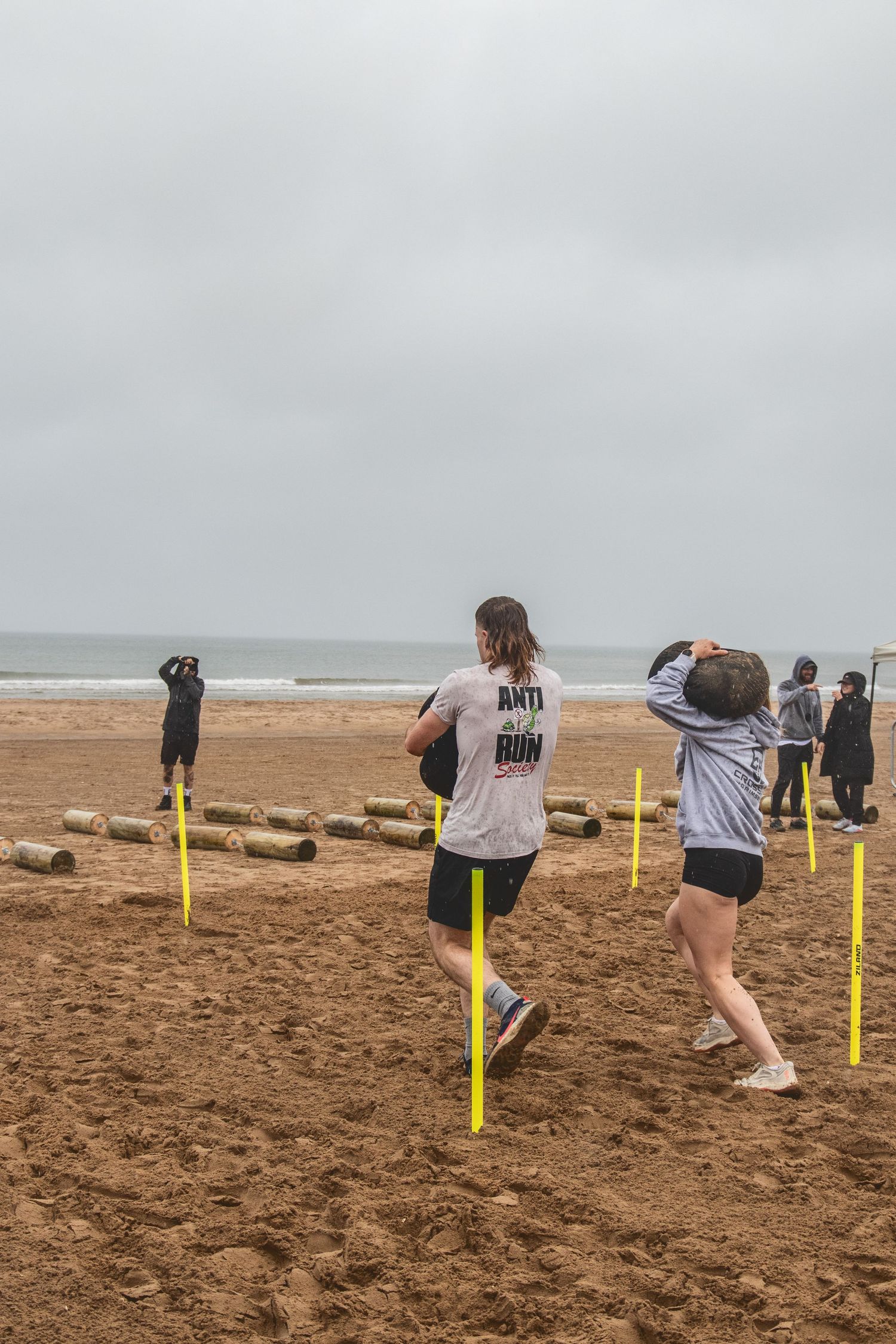 People participate in a beach fitness workout with yellow poles on sandy shore under overcast sky.