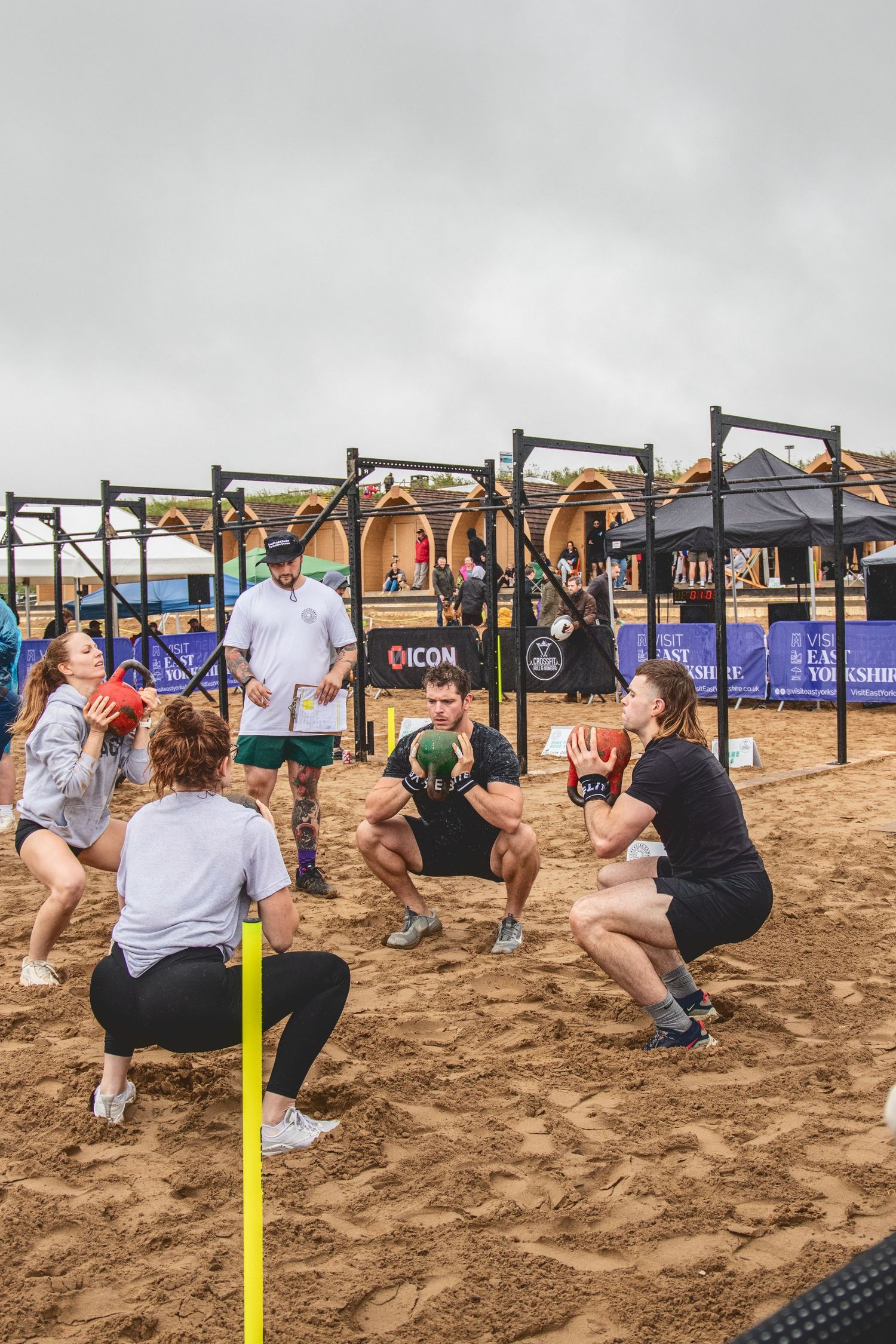 Athletes perform squats together during an outdoor CrossFit competition on a sandy beach with pull-up rigs in the background.