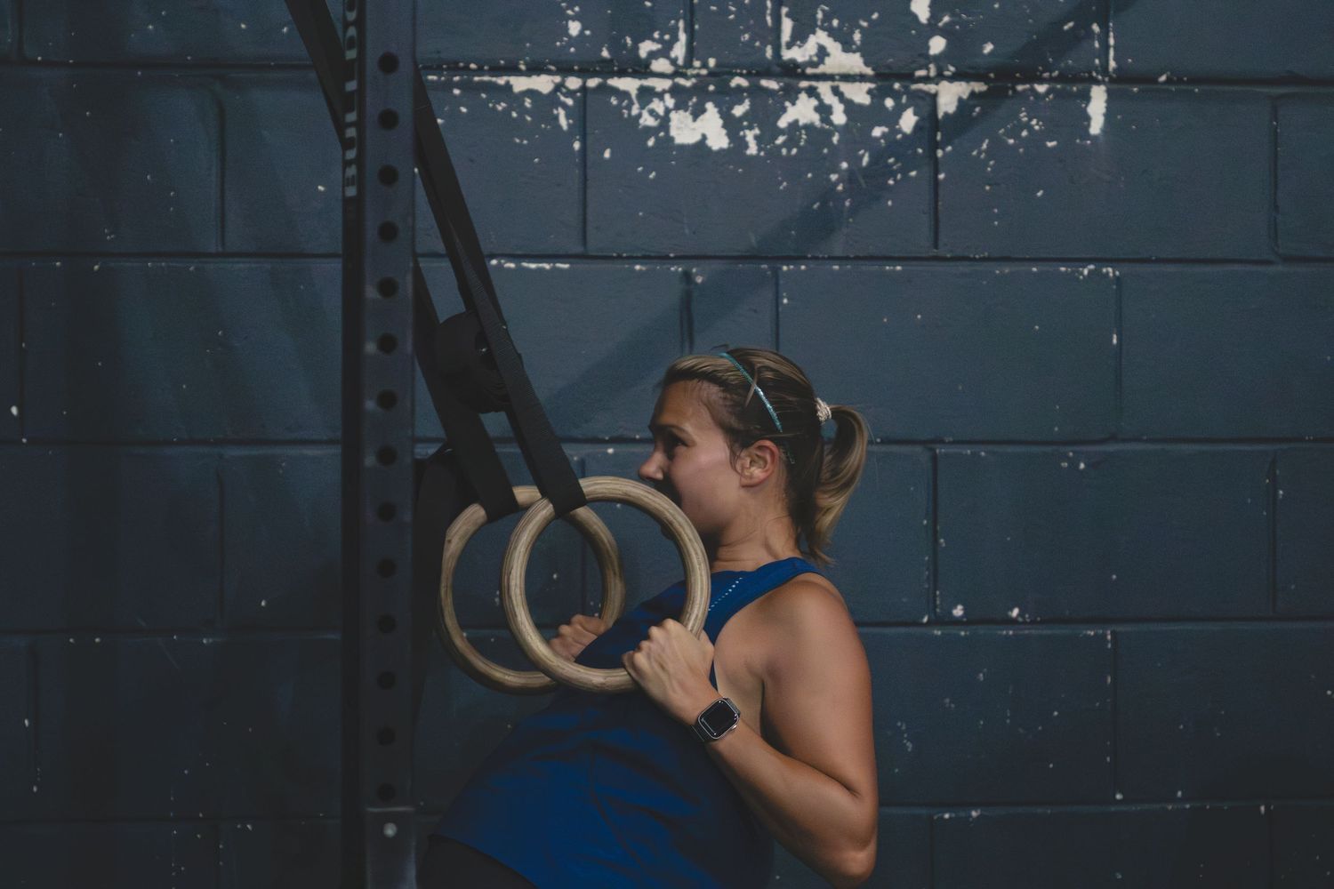 An athlete grips wooden gymnastic rings while training against a dark blue wall in a CrossFit gym setting.