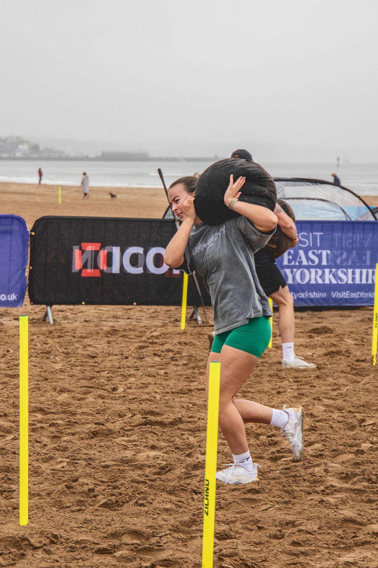Two athletes compete in a beach obstacle course event with yellow poles on sandy terrain near the ocean.