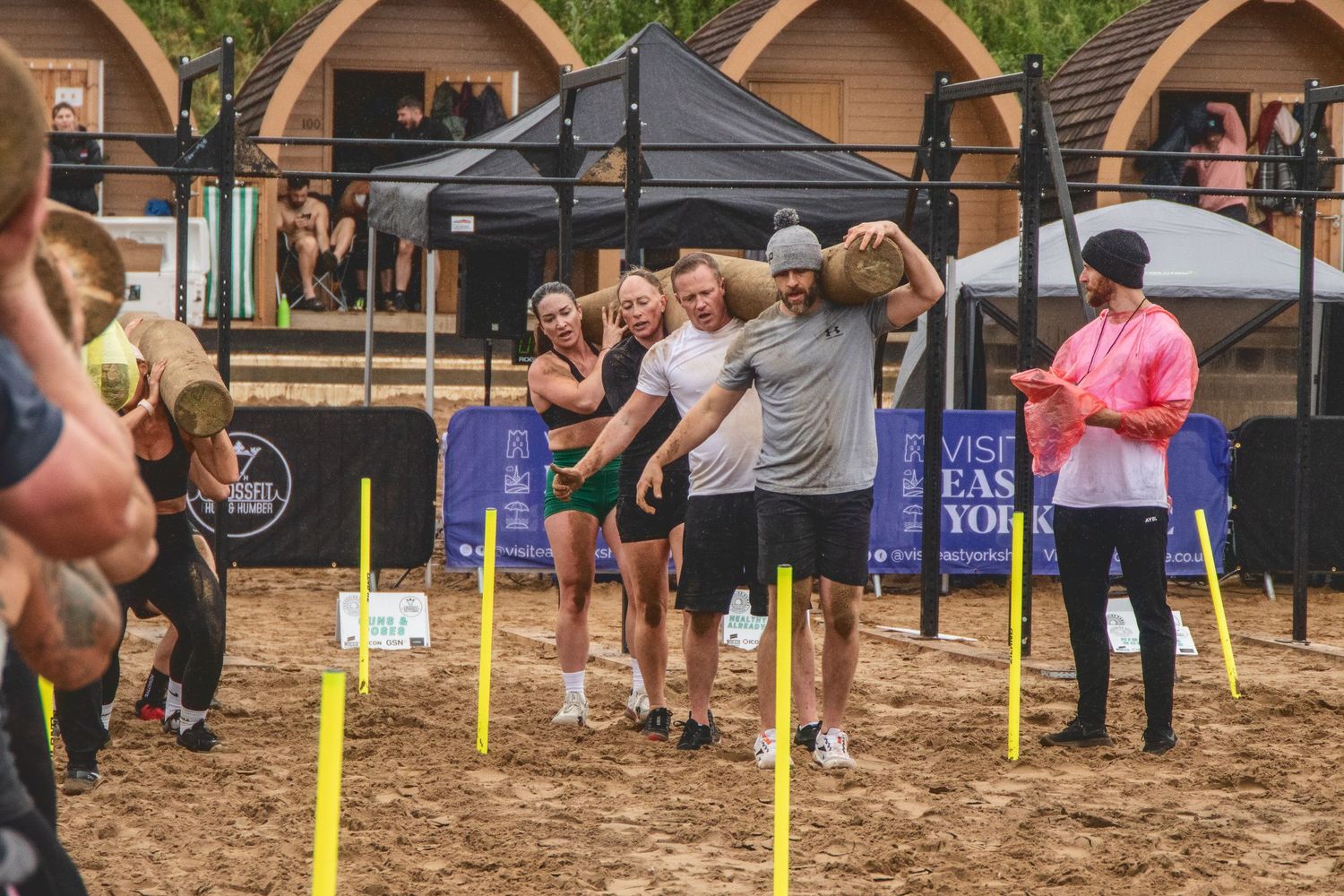Athletes compete in an outdoor fitness competition on dirt ground under canopy tents near wooden cabins.