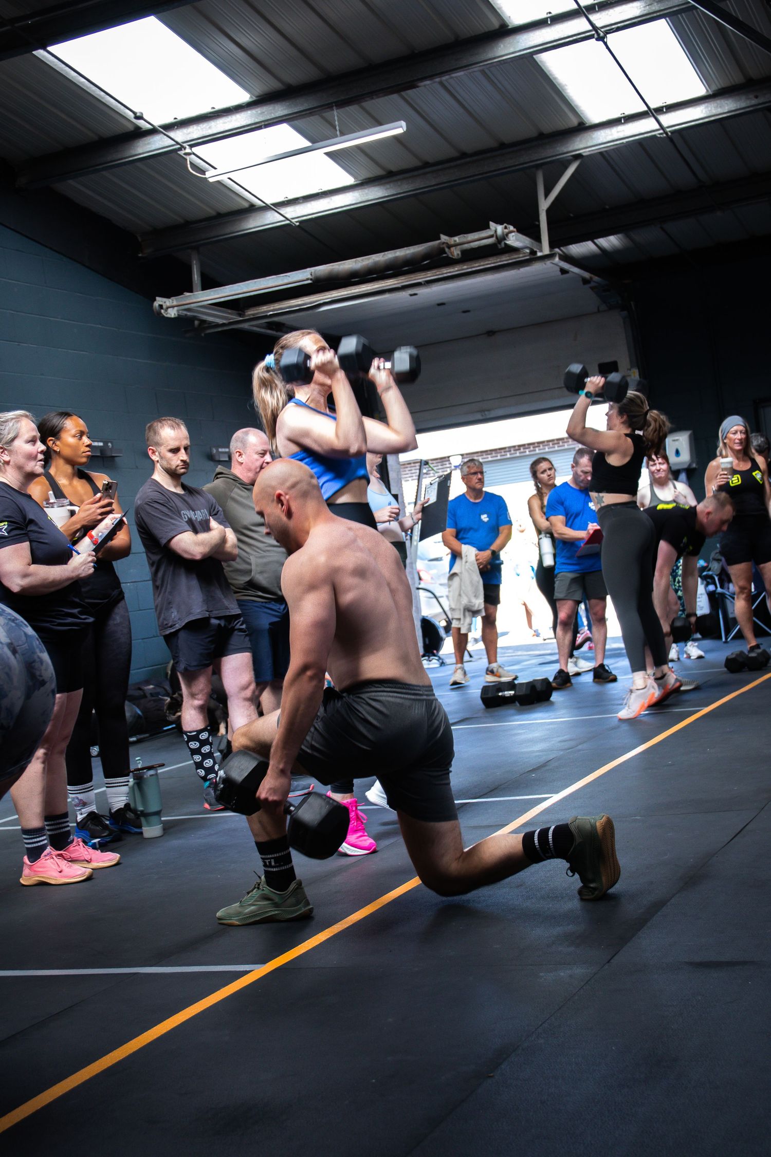 Athletes perform partner exercises with dumbbells during a CrossFit workout class as spectators watch from the sidelines.