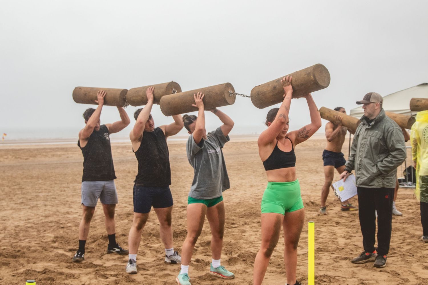 A group of people squatting in a line while holding logs overhead on a beach during team training exercise.