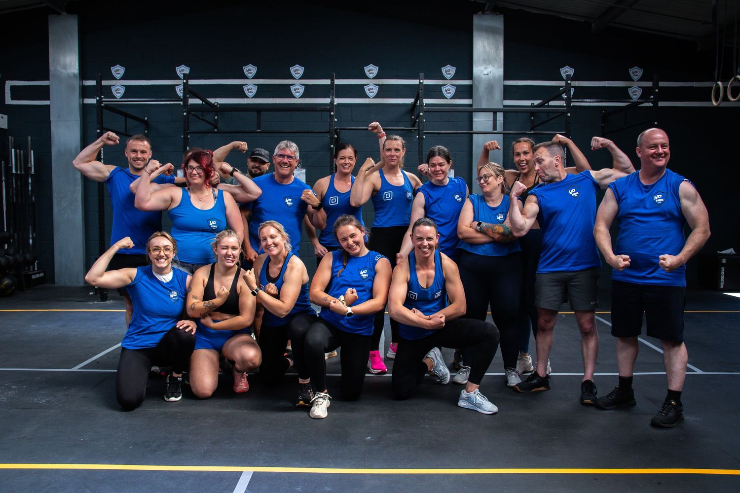 A group of fitness enthusiasts in matching blue tank tops pose together at a gym facility.
