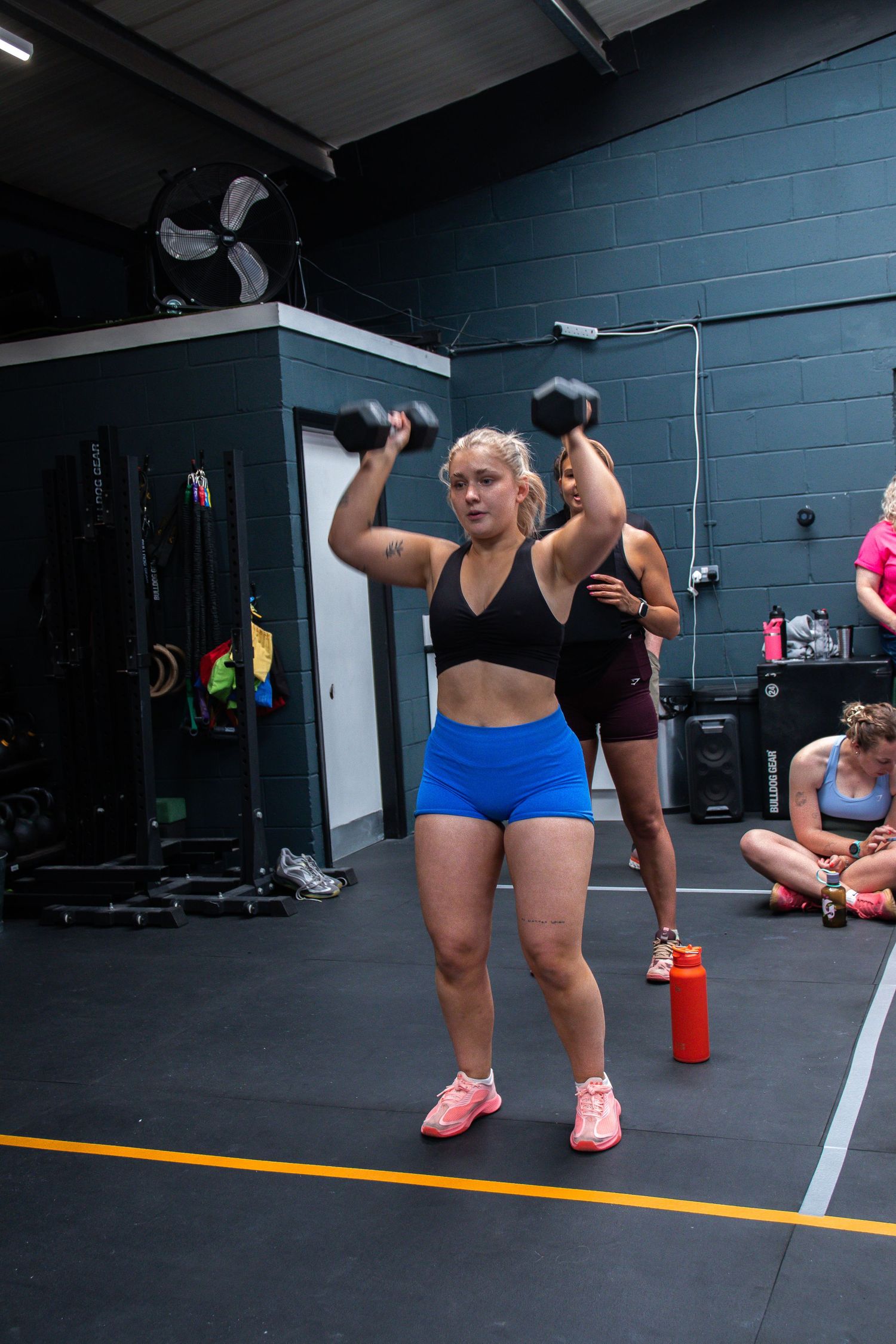 A person in a black sports bra and blue shorts performs dumbbell exercises in a CrossFit-style gym facility.