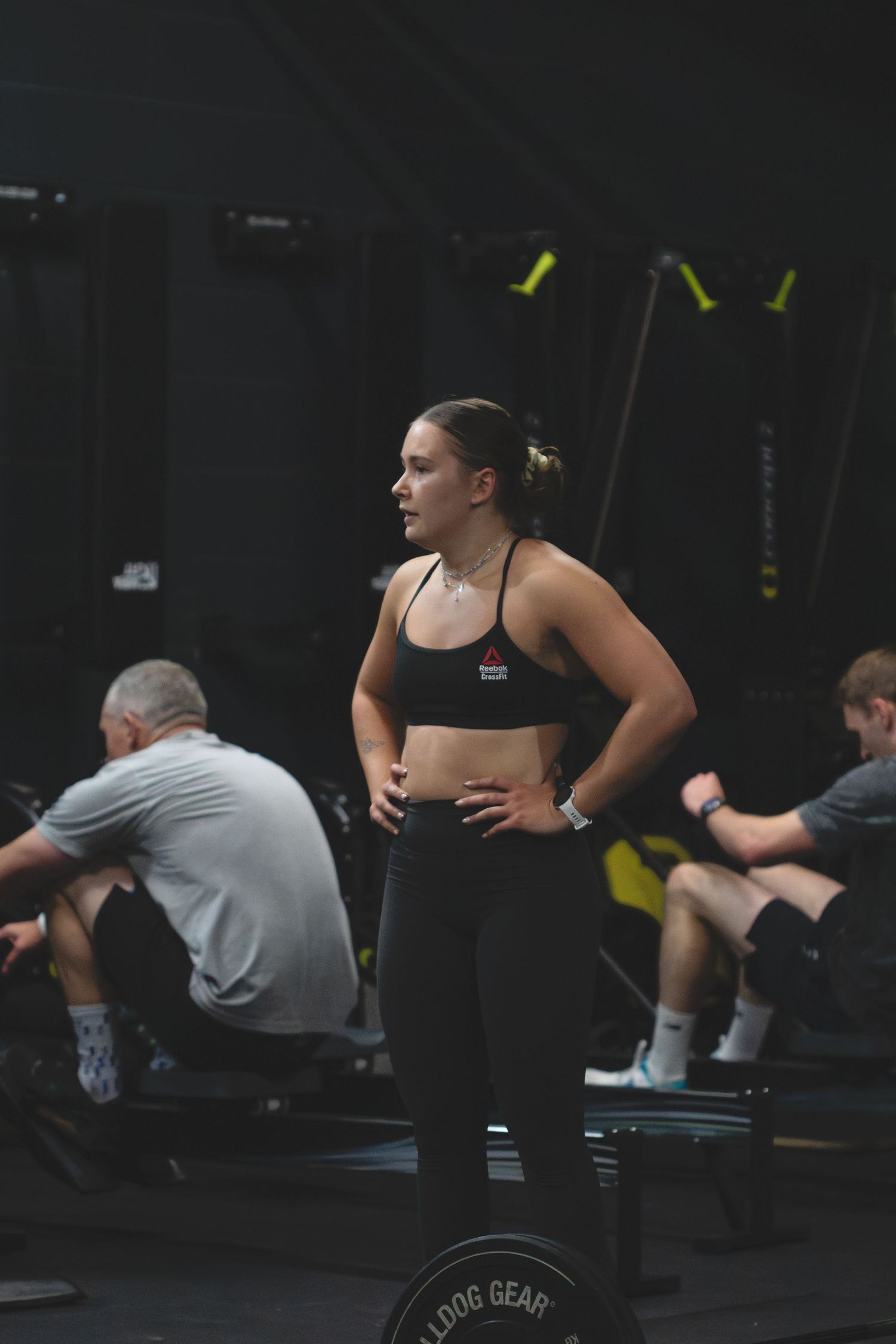 An athlete in black workout attire takes a break between exercises at a dimly lit gym with weightlifting equipment.