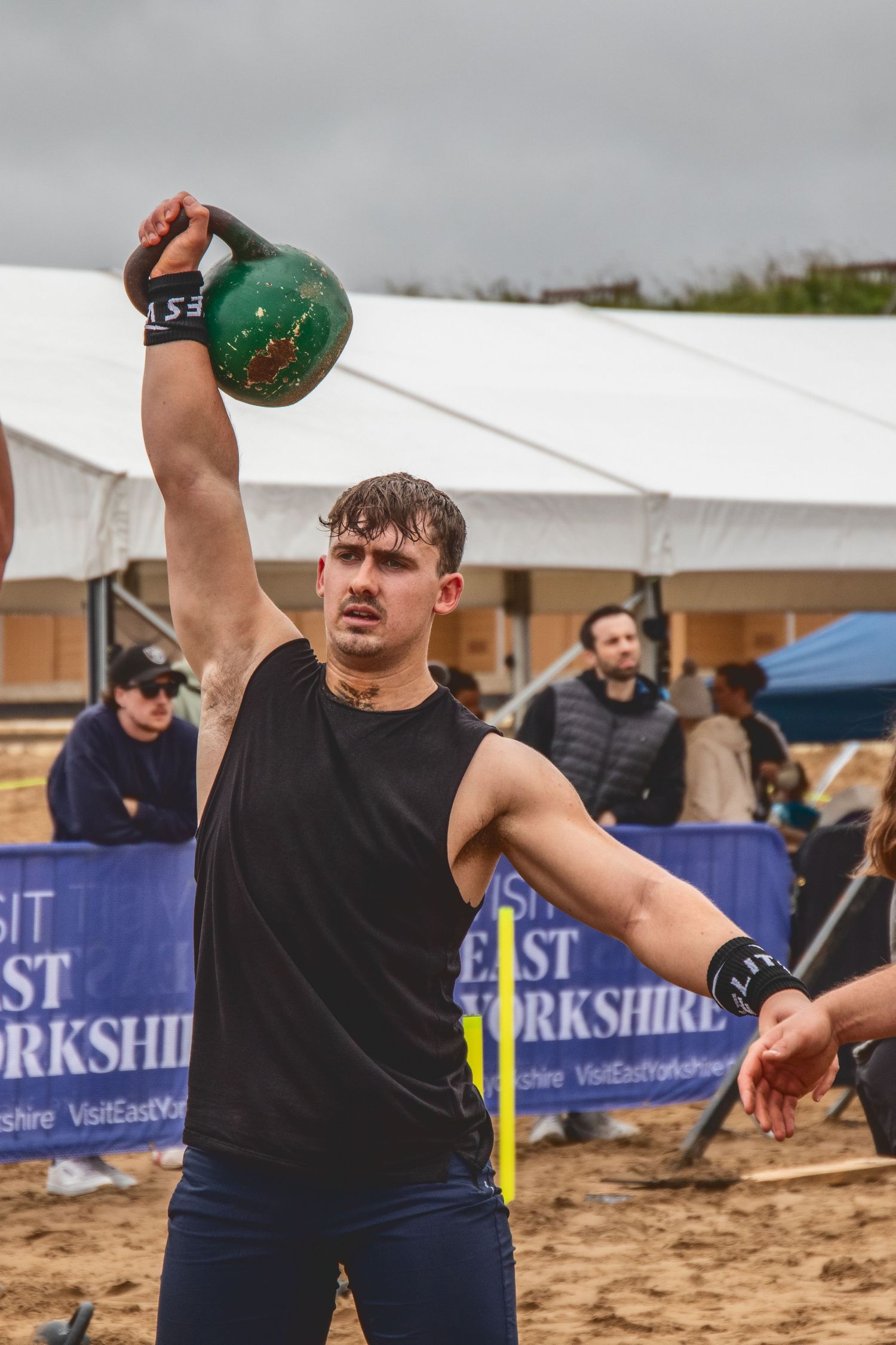 Athlete performs kettlebell lift during outdoor competition on sandy beach with white tent in background.