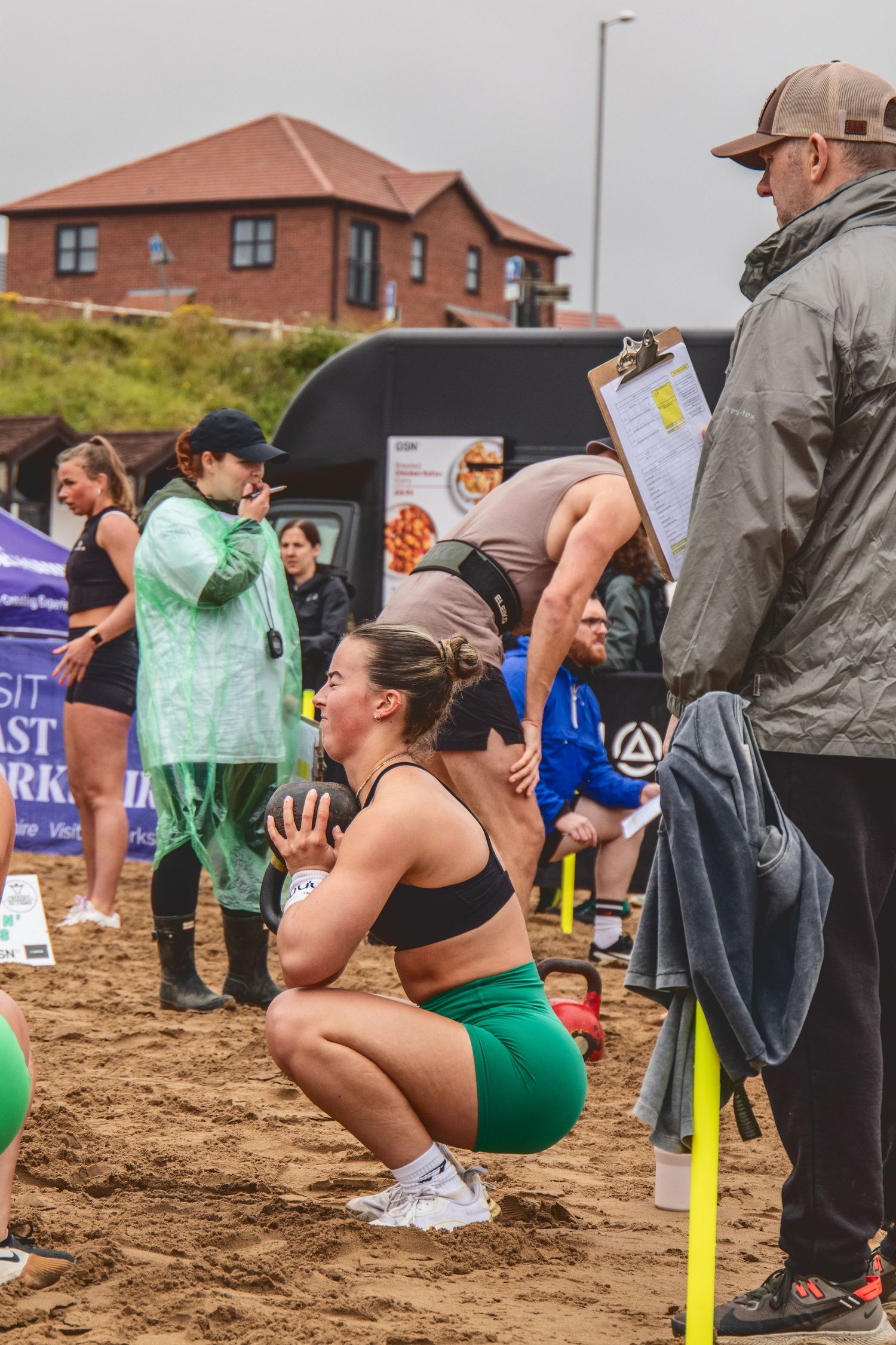 Athletes compete in an outdoor sandbag lifting and carrying competition on muddy terrain near brick buildings.