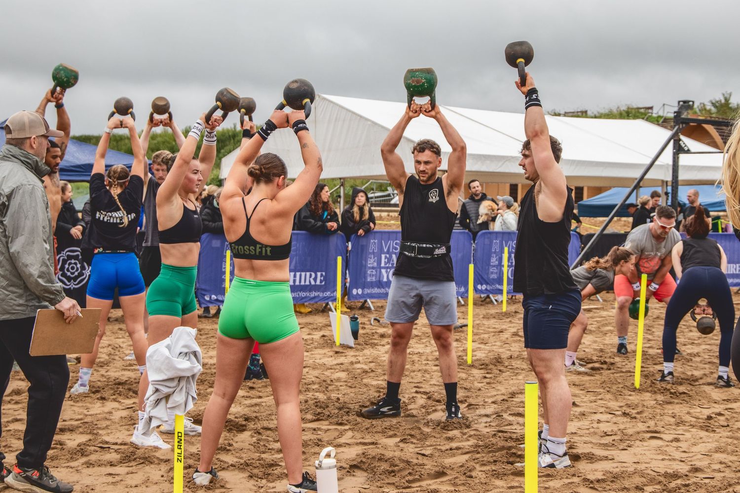 Athletes perform synchronized kettlebell exercises on a sandy beach during an outdoor fitness competition event.