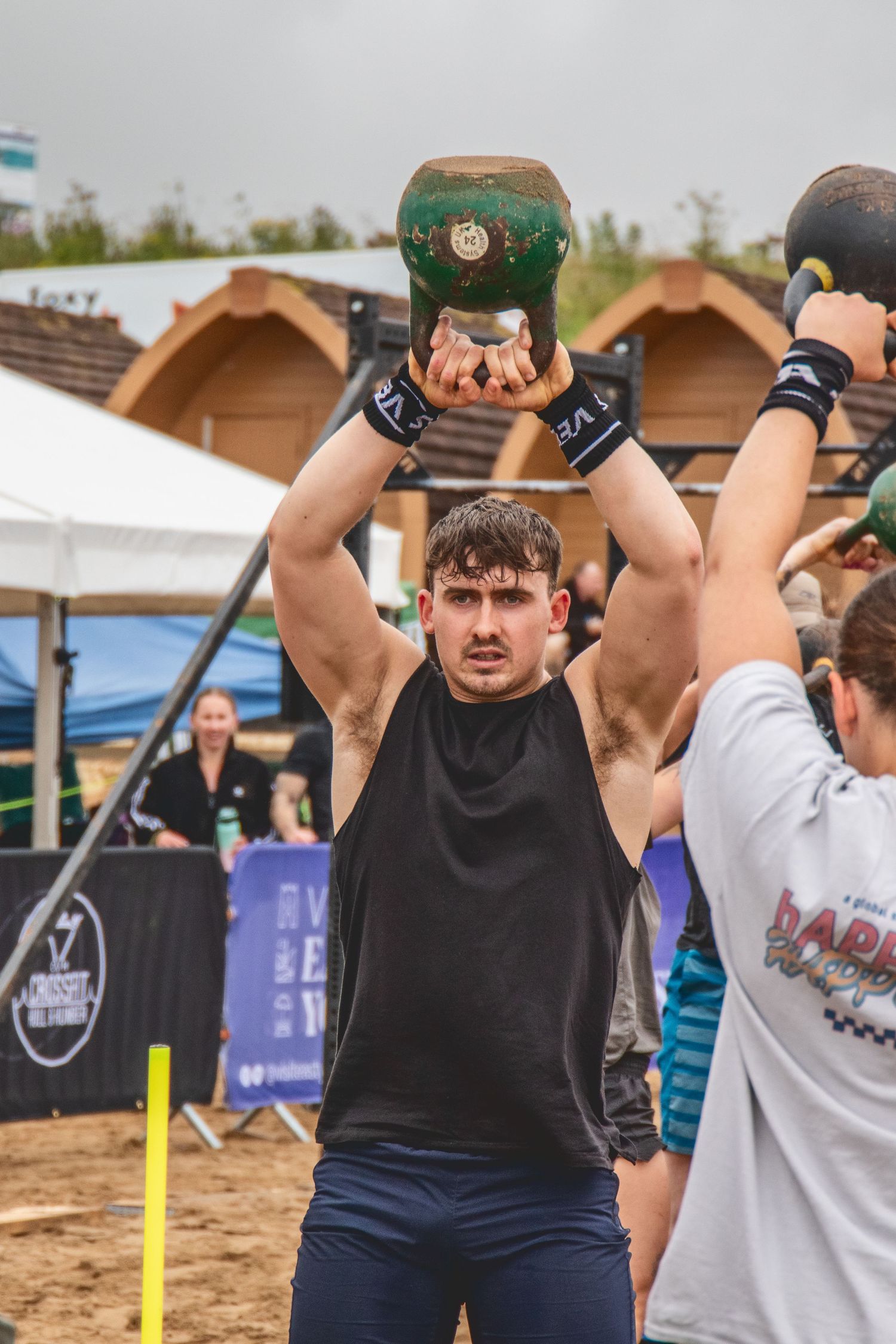 An athlete in a black tank top performs an overhead kettlebell lift during an outdoor fitness competition.