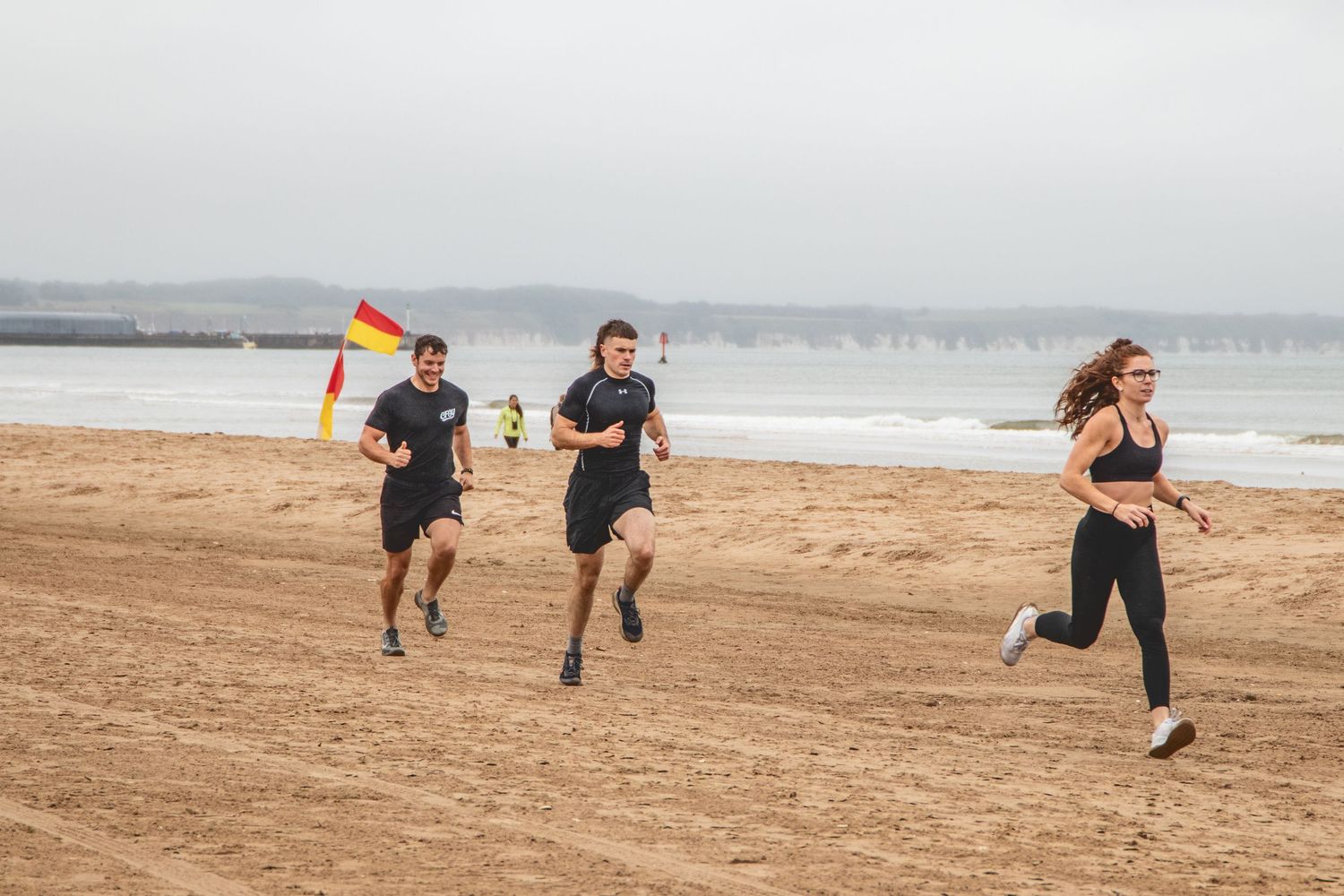 Runners training on sandy beach on cloudy day with ocean waves in background.