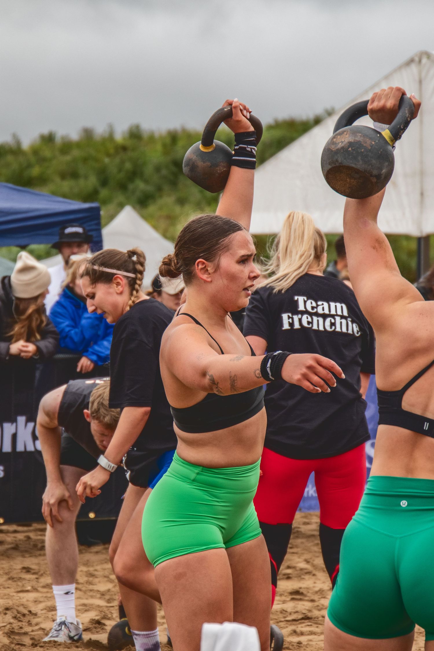 Athletes perform kettlebell lifts during an outdoor CrossFit competition while wearing colorful athletic gear.