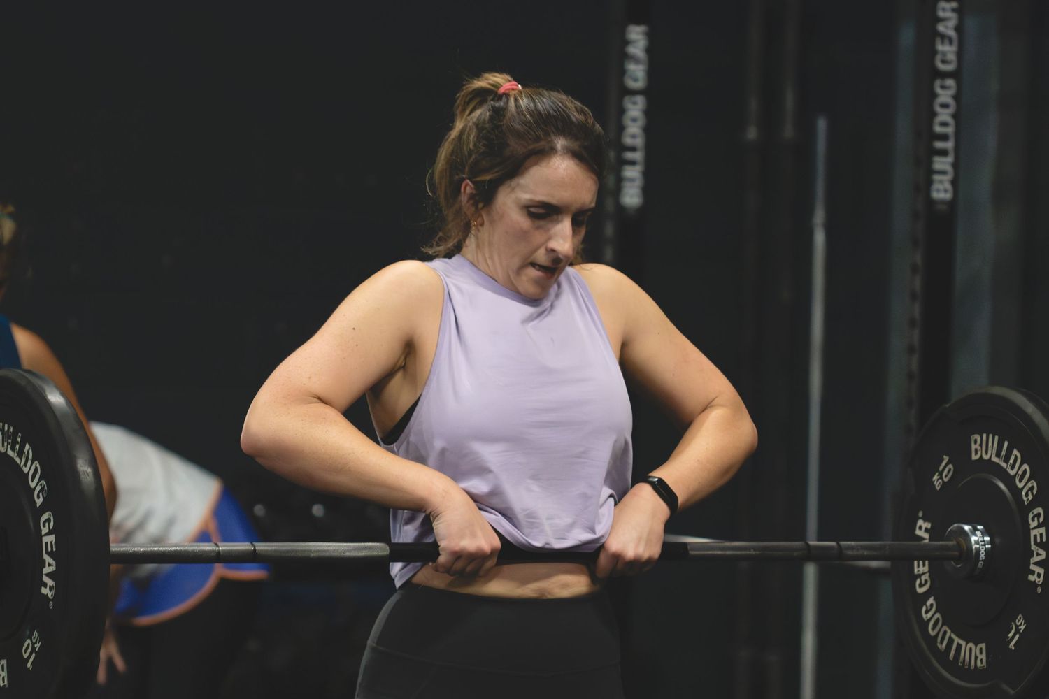 An athlete in a light purple tank top adjusts their weightlifting belt while preparing for a heavy barbell lift at the gym.