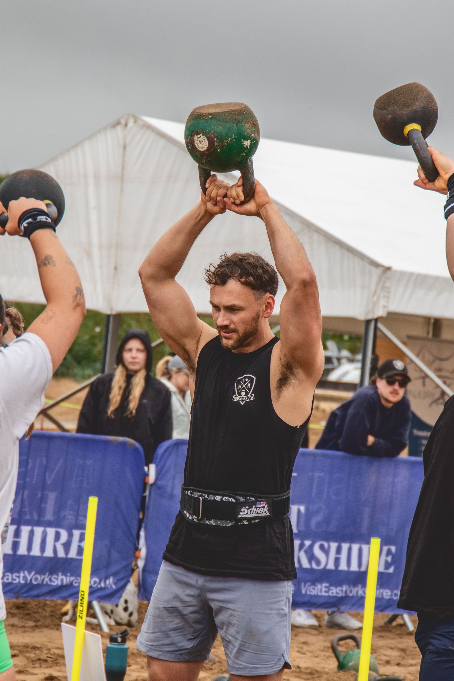 Athlete competing in strongman competition lifting heavy kettlebells overhead at an outdoor event with crowd barriers.