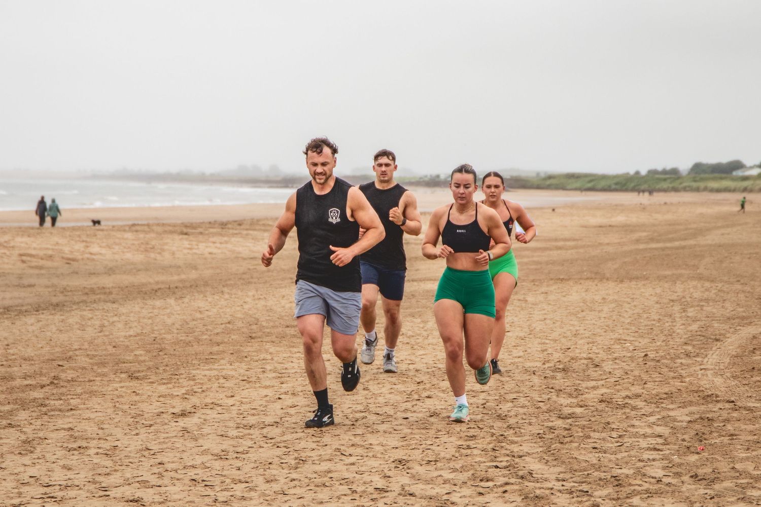 Athletes run together on a grey sandy beach during group outdoor fitness training on an overcast day.