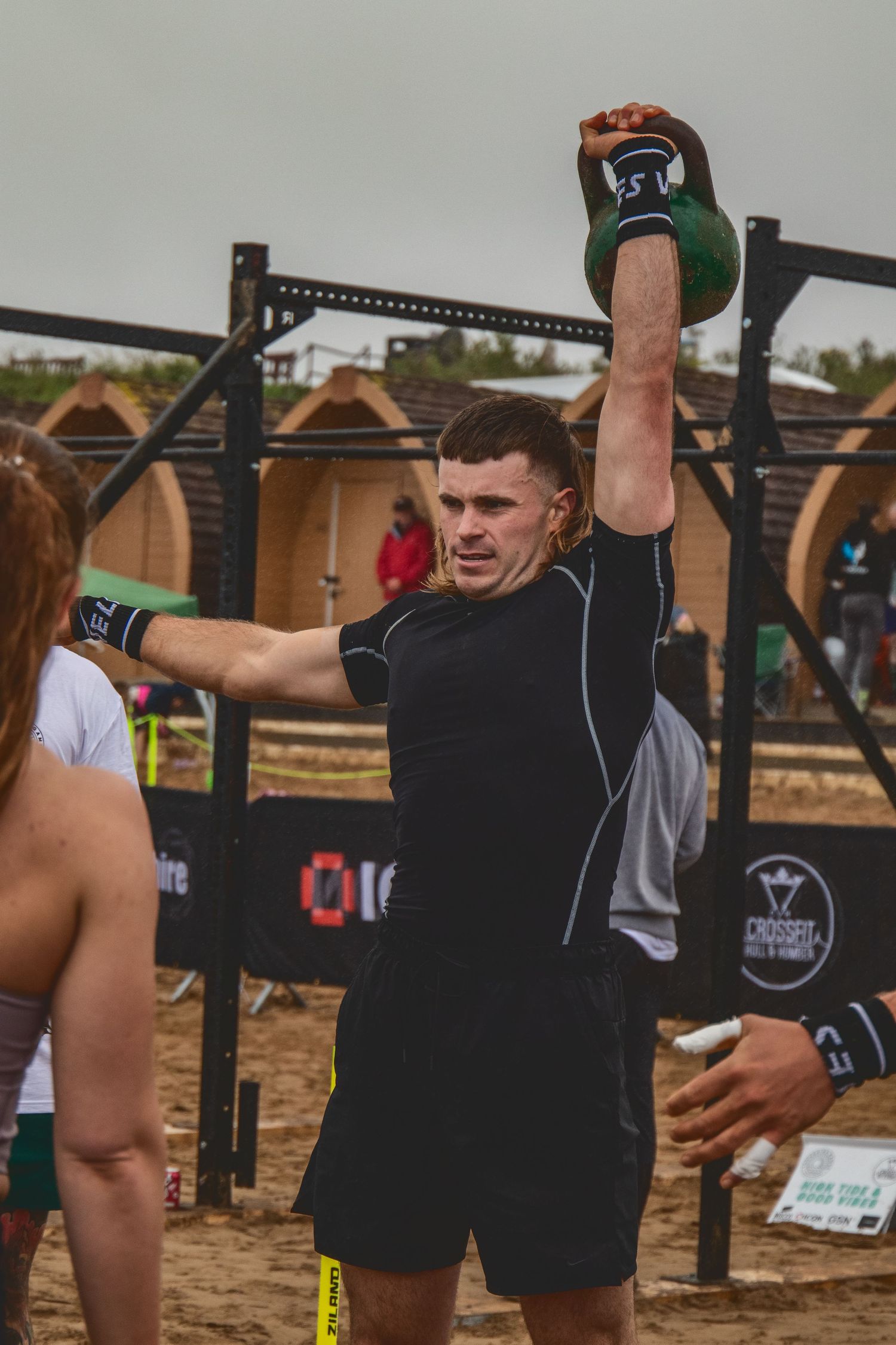 Athletes engaged in an outdoor CrossFit competition performing exercises on pull-up bars in sandy terrain.