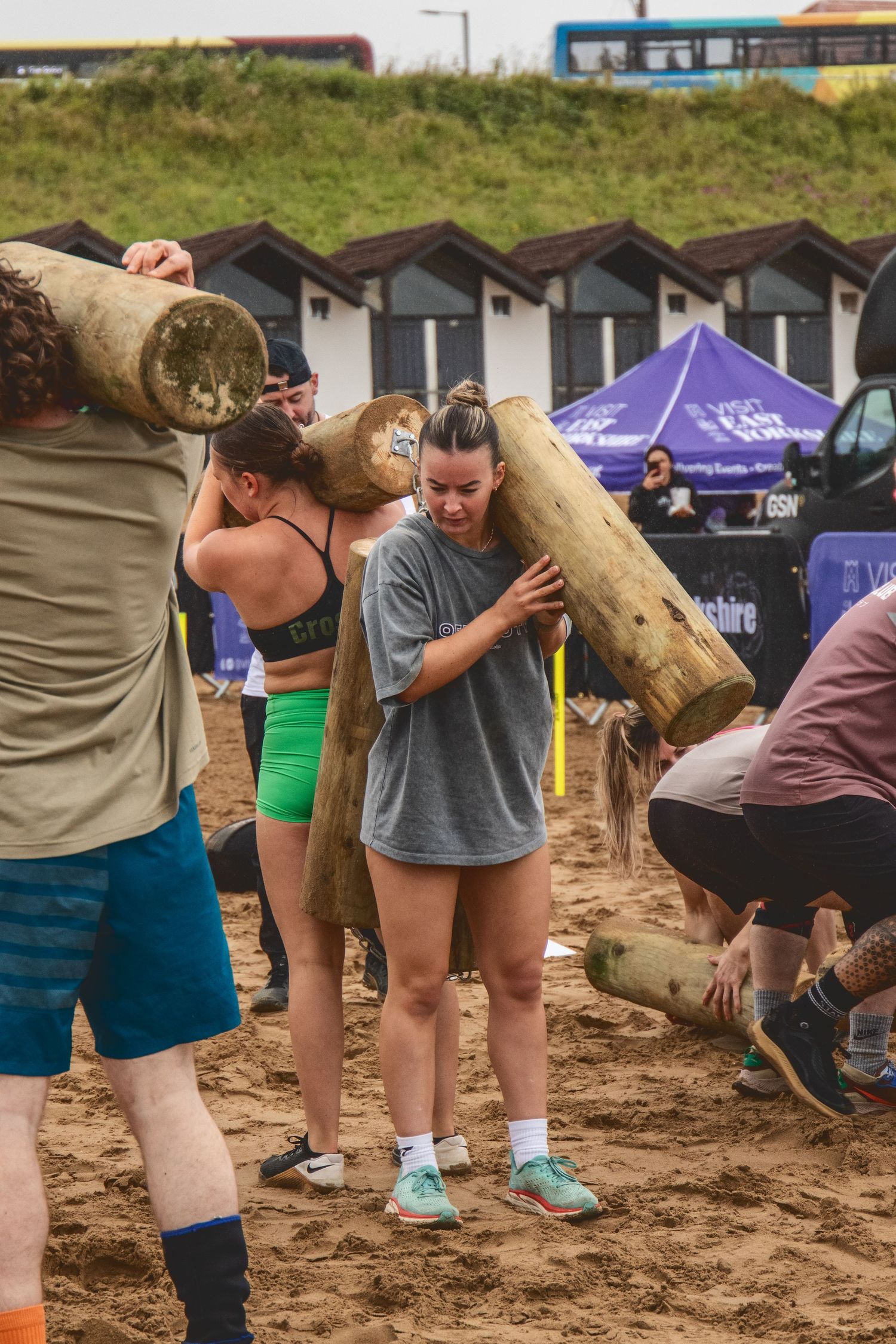 Competitors carry heavy wooden logs during an outdoor strength competition on sandy beach terrain near beach huts.