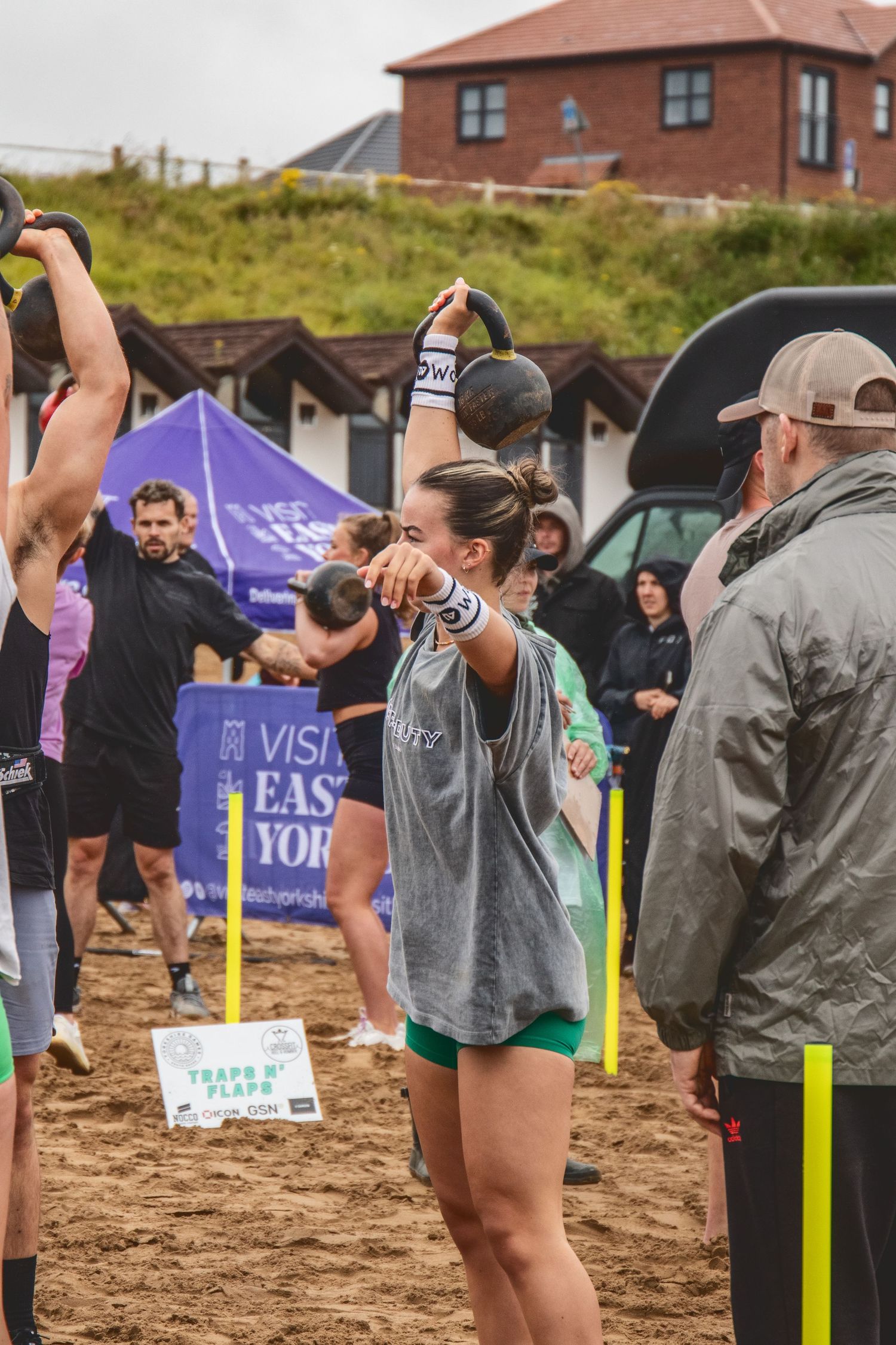 Athletes compete in an outdoor fitness competition on sandy terrain with exercise equipment and spectators gathered around.