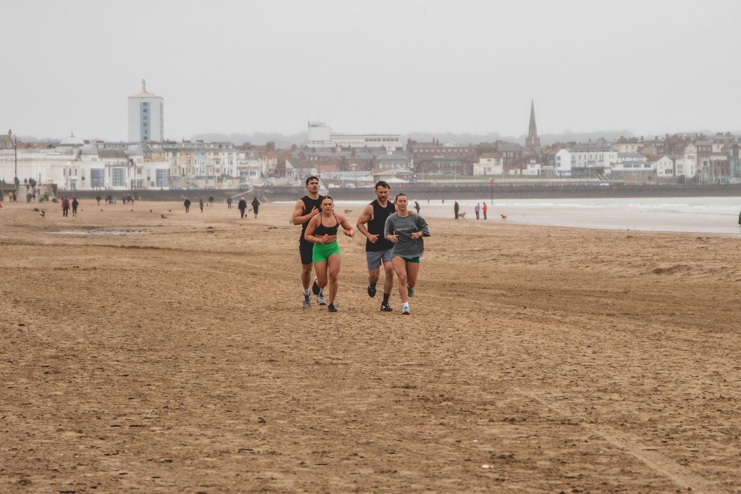 Three people jog together on a sandy beach with a coastal city skyline visible in the background.