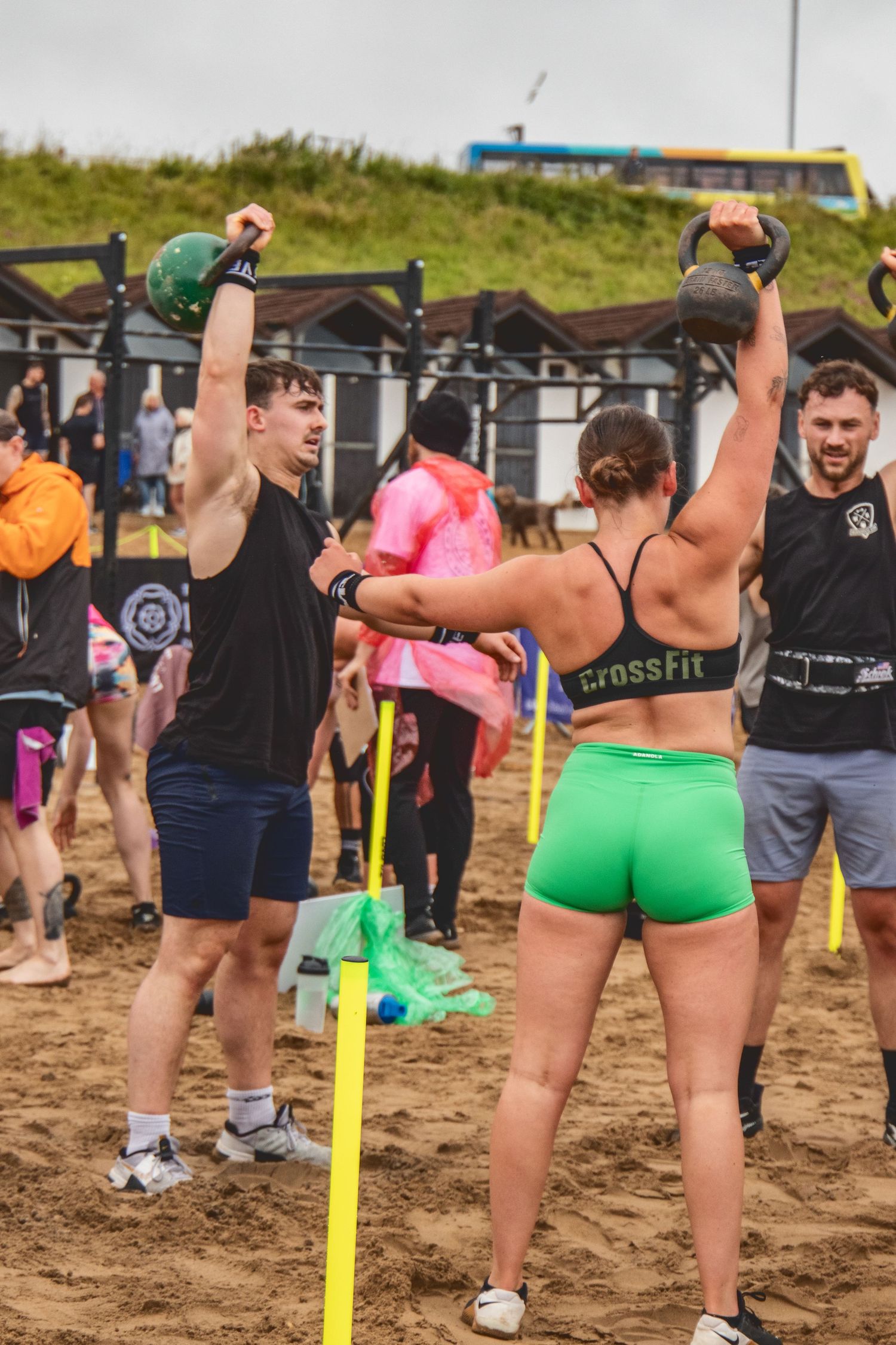 Athletes perform outdoor CrossFit exercises with kettlebells on a sandy beach during a fitness competition.