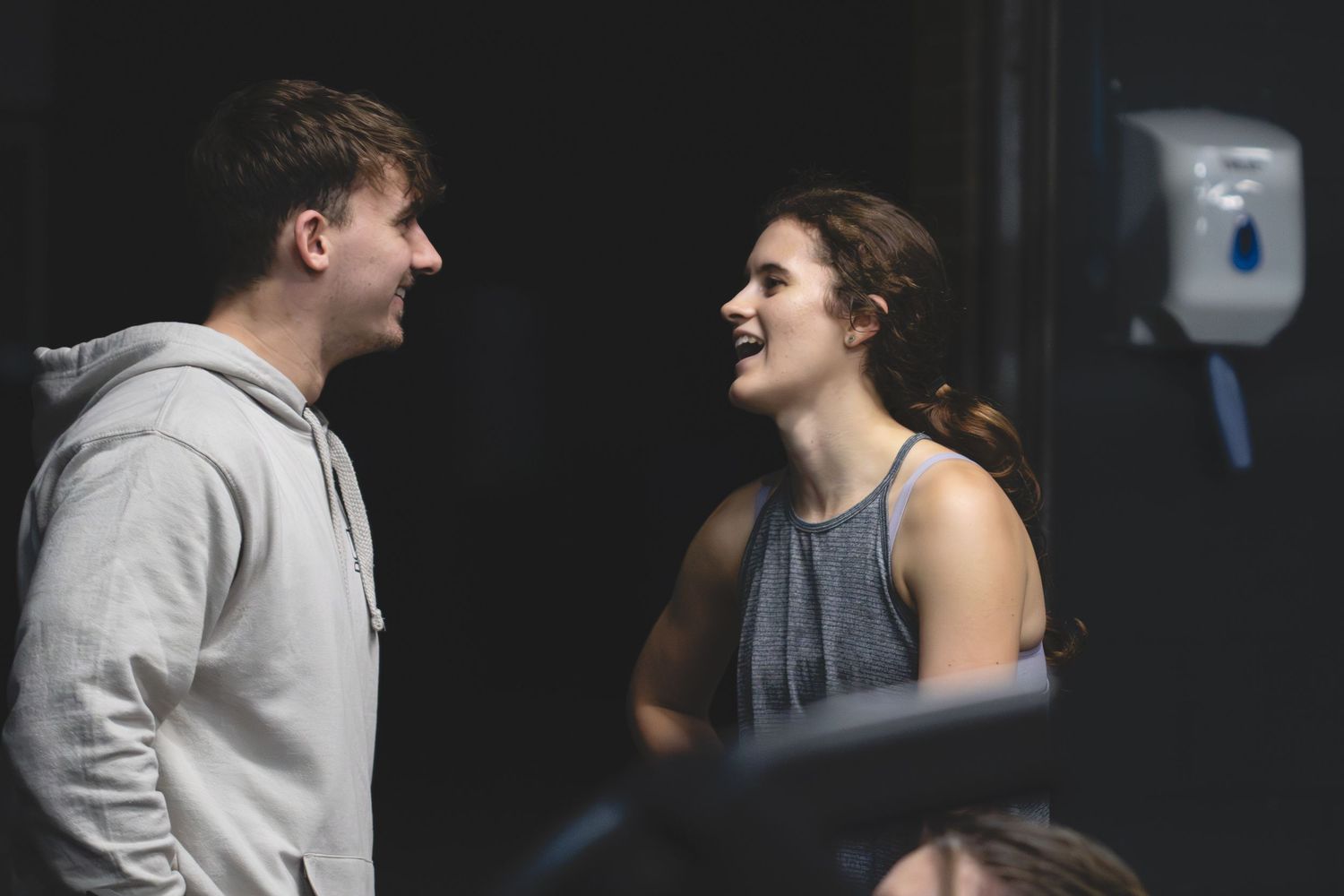 Two people laughing and engaging in animated conversation while standing in a dimly lit indoor setting.