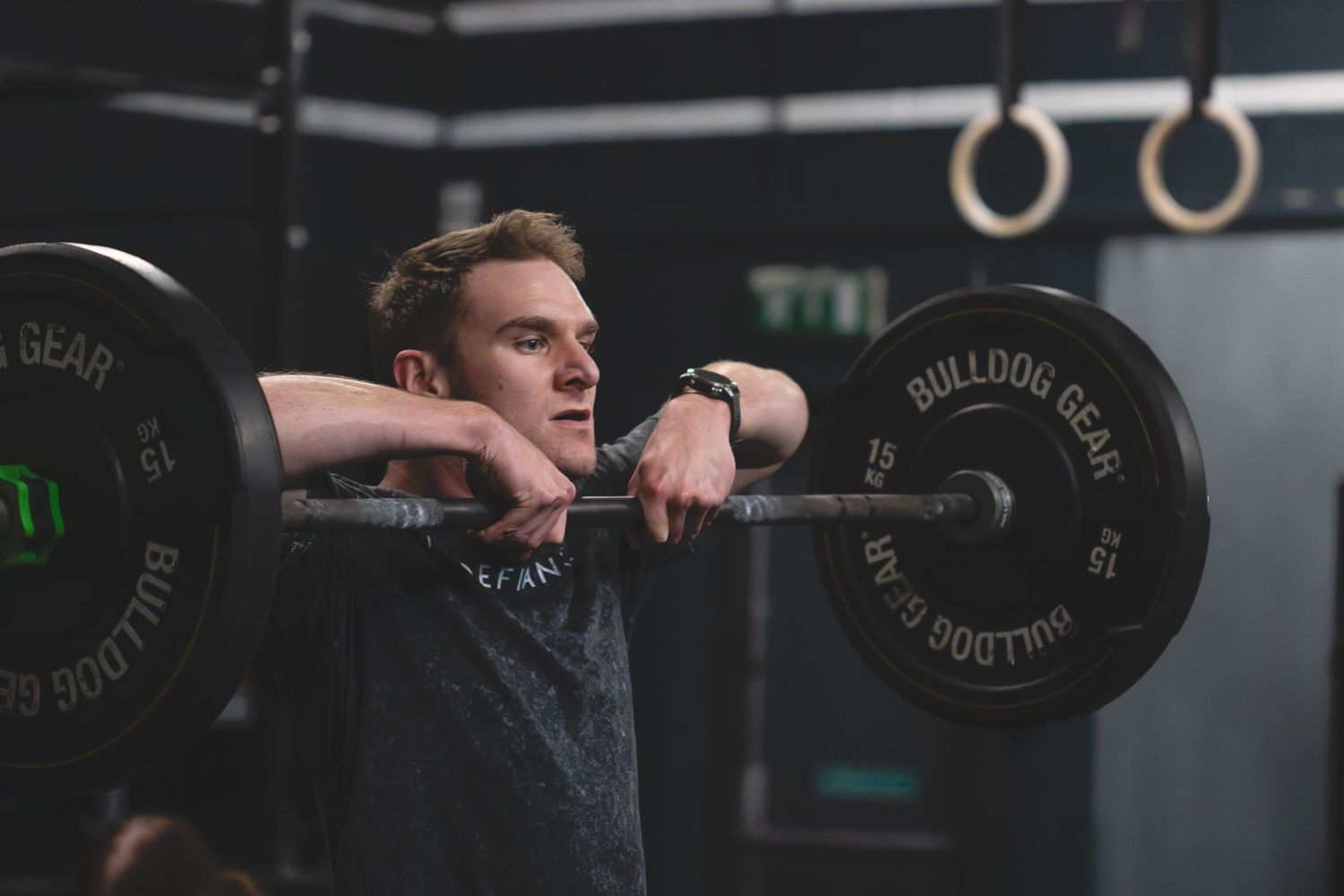 An athlete rests on a heavy barbell during a workout in a dark gym with gymnastic rings visible in the background.