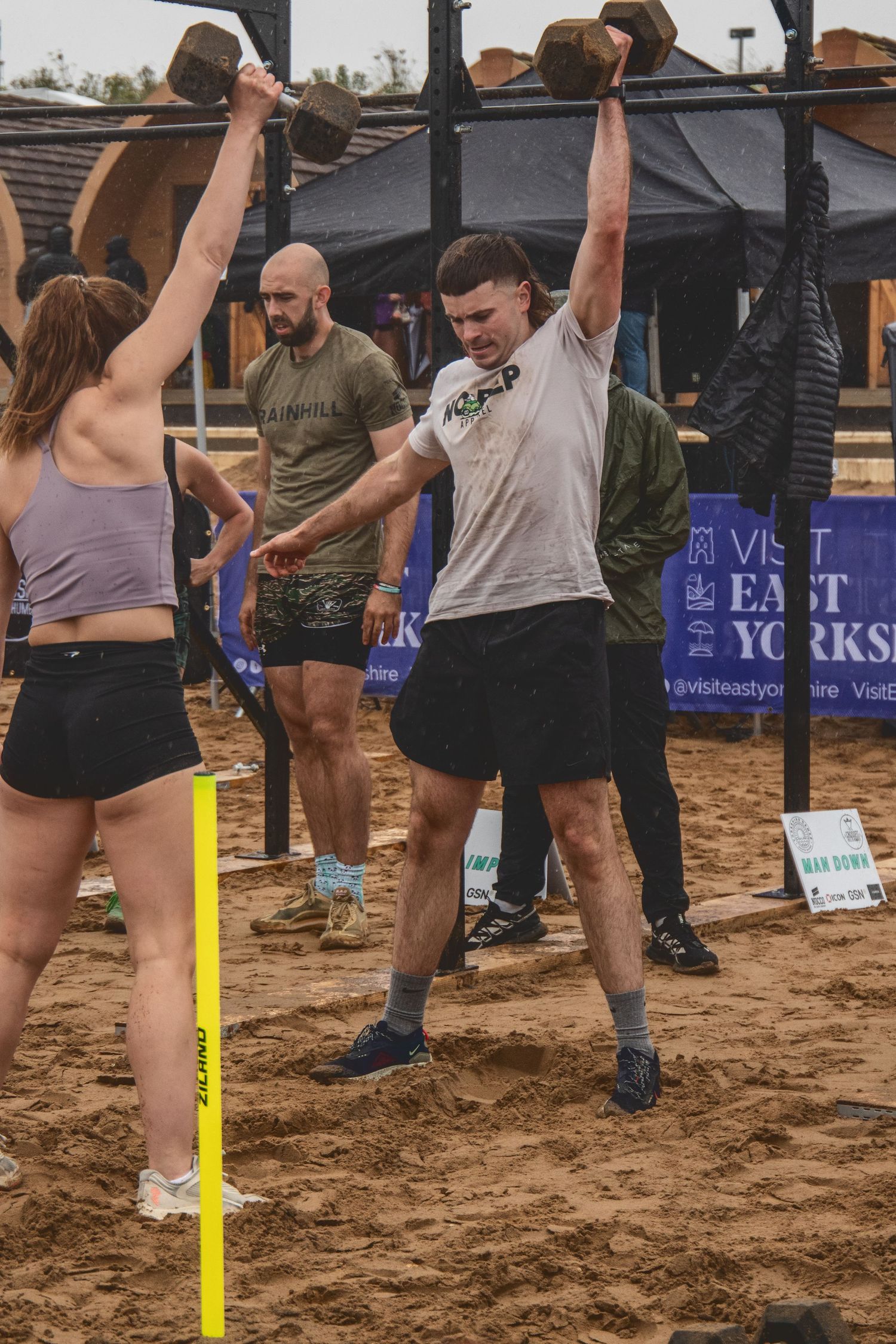 Athletes perform overhead dumbbell exercises during an outdoor fitness competition on sandy ground.