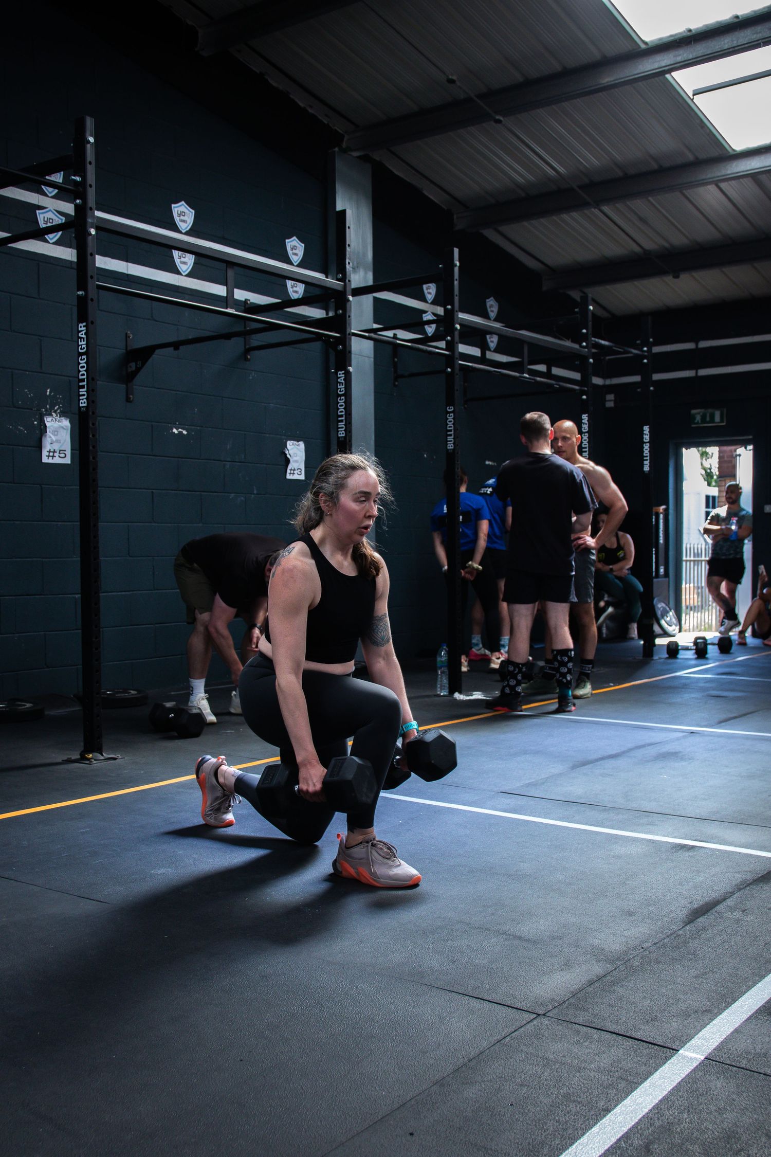 A person performs lunges with dumbbells during a workout session in a dark CrossFit-style gym facility.