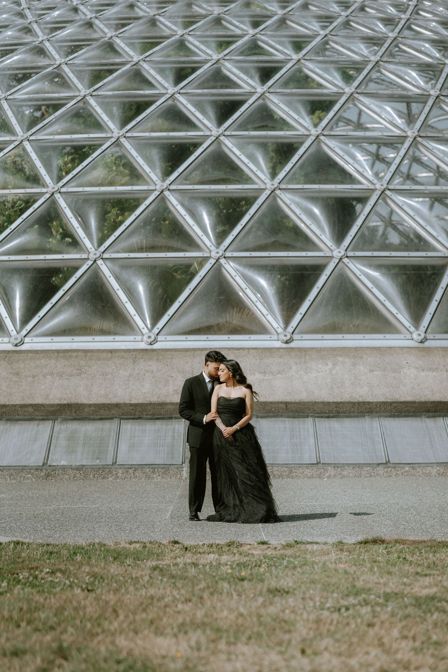 Architectural detail showing a geometric metal grid structure with two figures sitting beneath it on grass.