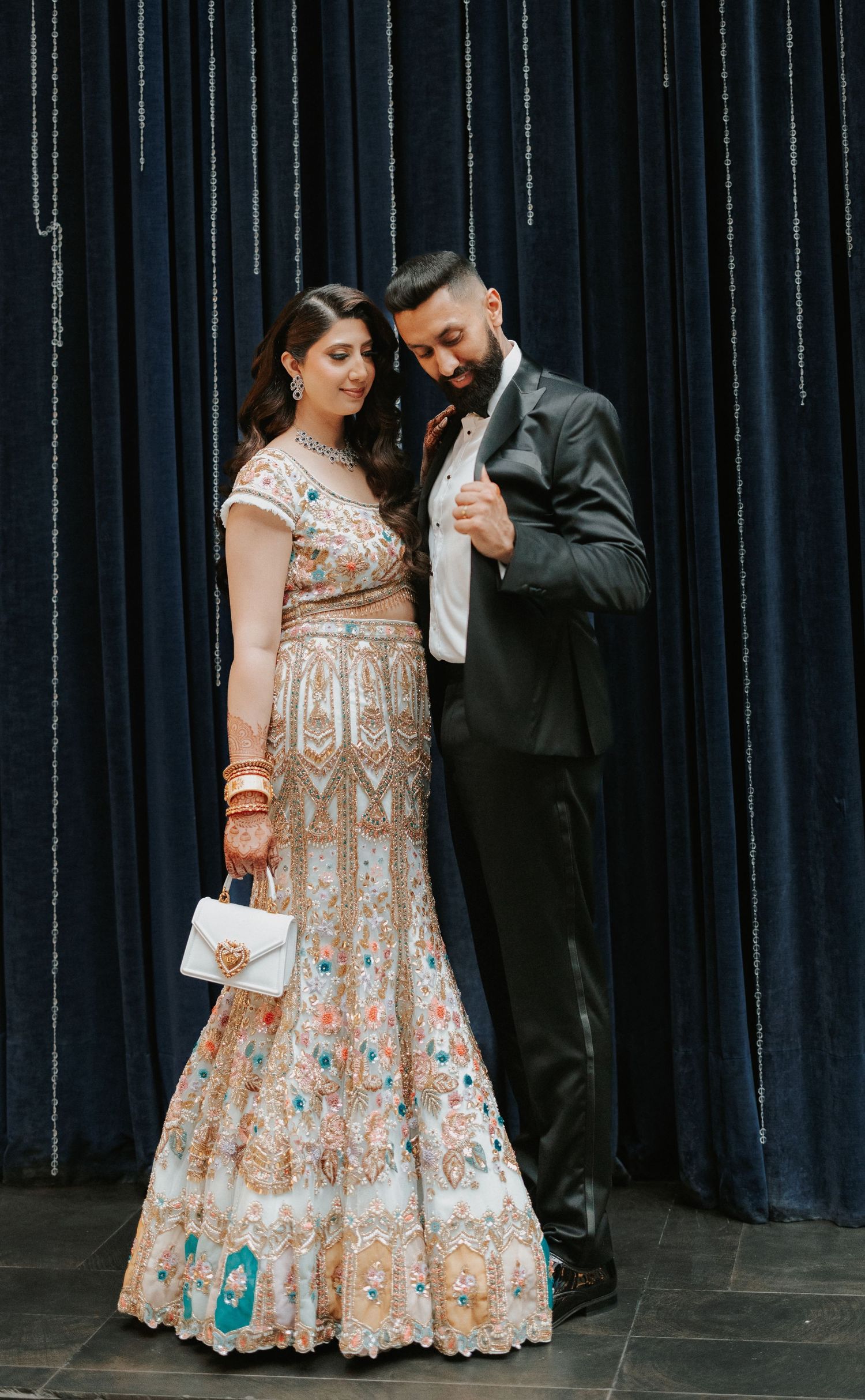 Romantic couple poses against dark backdrop with bride in ornate beige and turquoise lehenga dress.