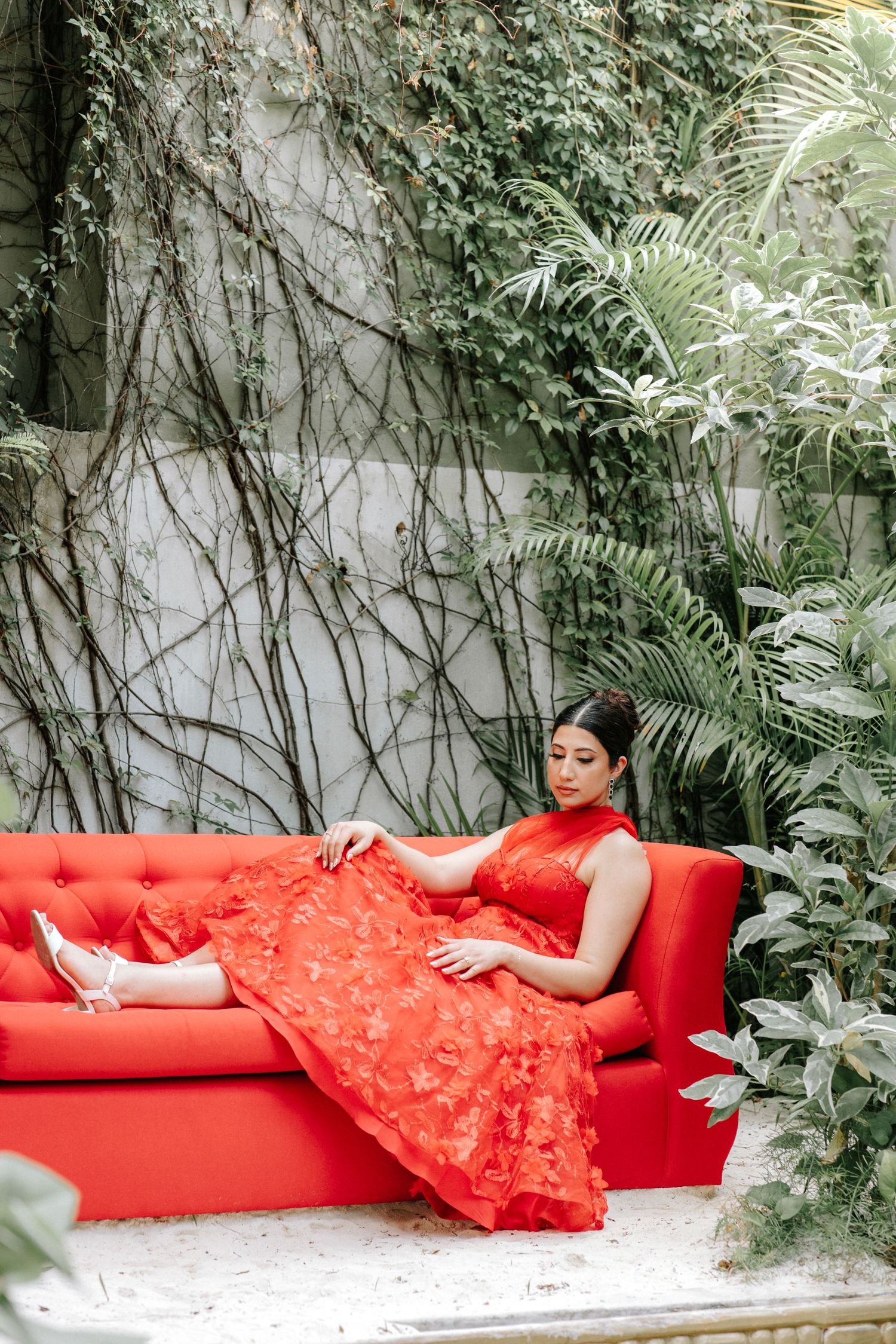 A figure in a flowing red lace dress reclines on a red chaise lounge against a backdrop of lush greenery and vines.