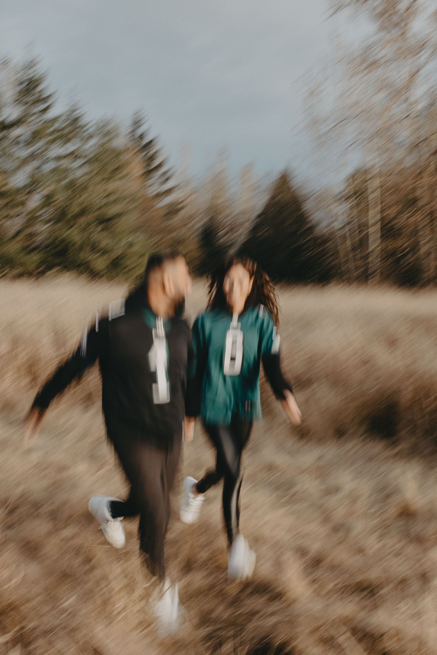 Two people in sports jerseys running together through a golden field with evergreen trees in the background.