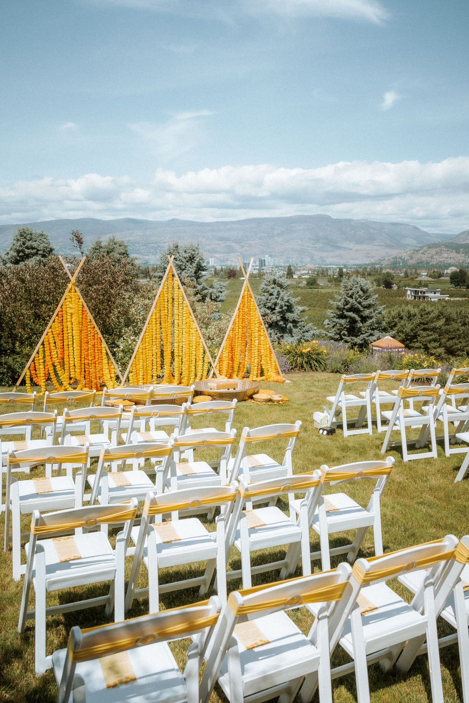 Outdoor wedding ceremony setup with white chairs and yellow teepee-style decorations against a mountain landscape backdrop.