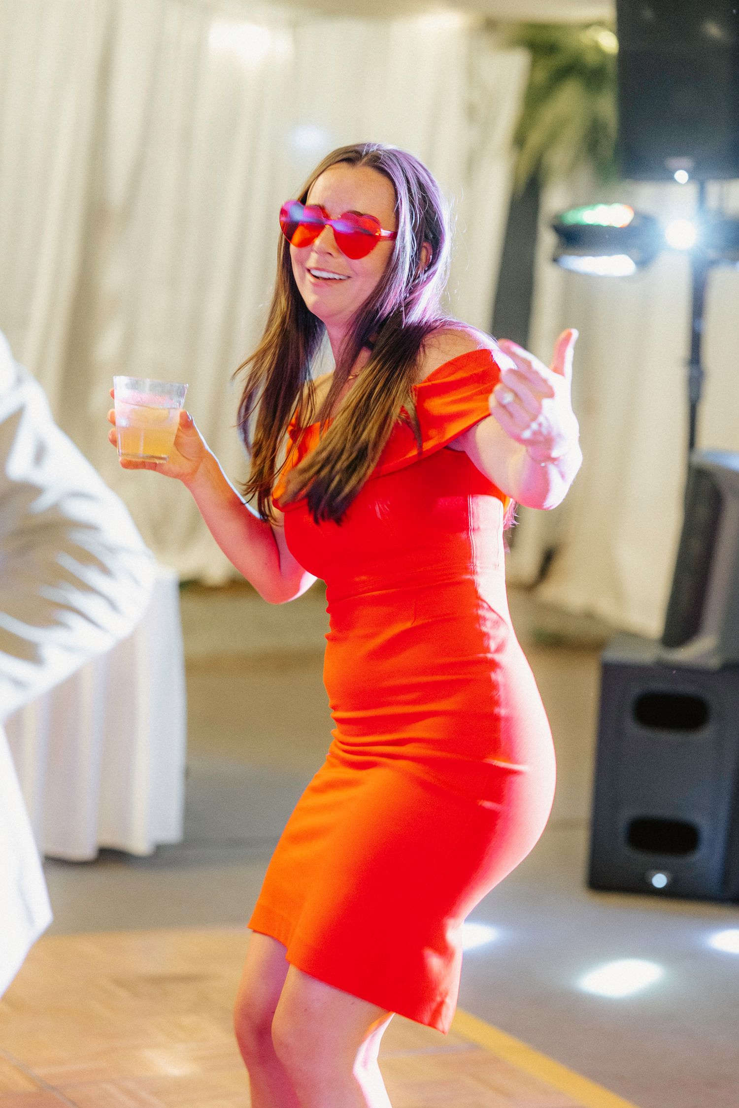 Wedding guest in bright orange dress dances at reception with sunglasses.