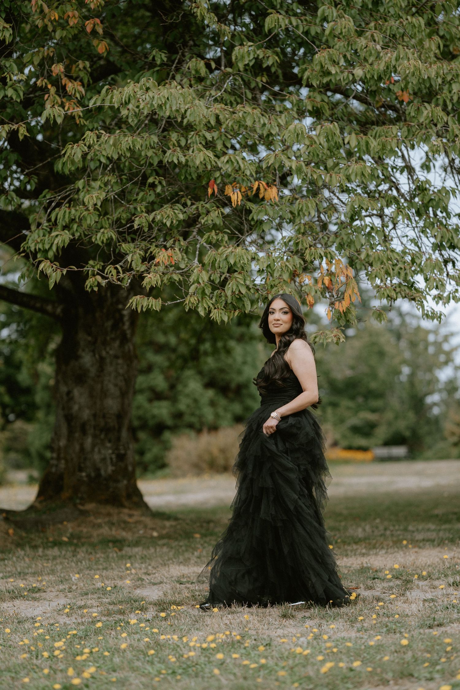 A person in an elegant black tulle gown poses gracefully under a large tree with autumn leaves falling in a park setting.