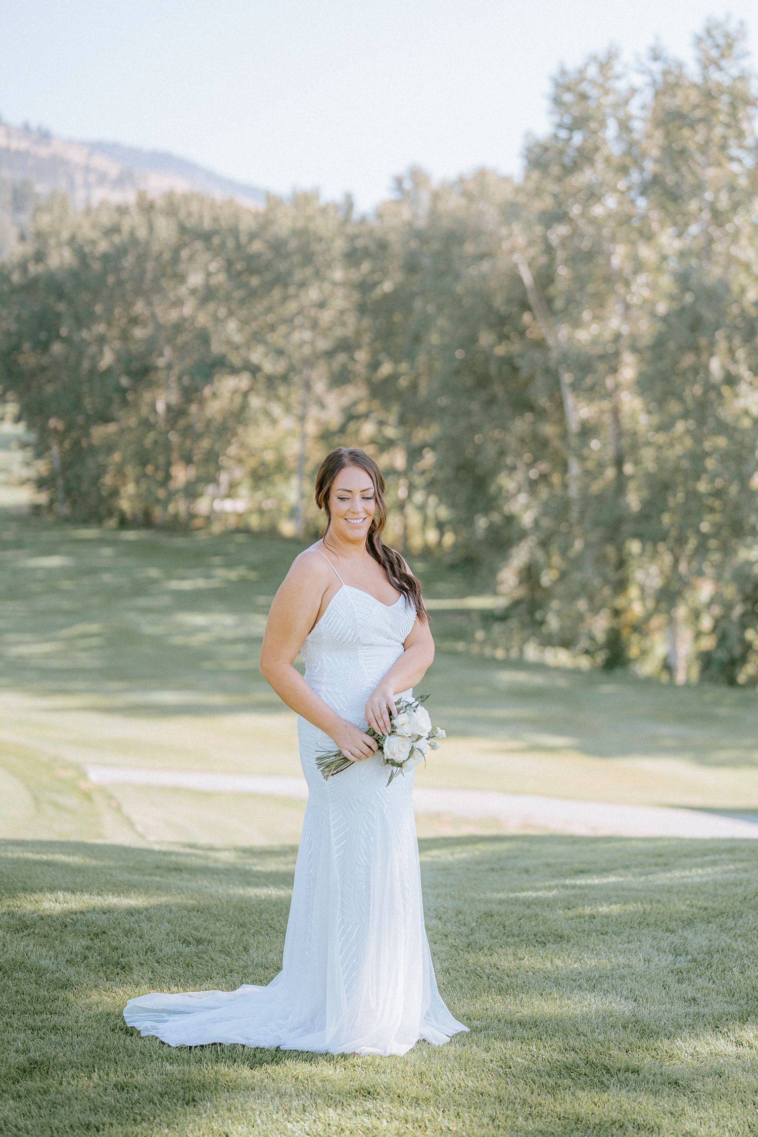 A bride in a white wedding dress poses outdoors on a grassy hillside surrounded by trees on a sunny day.