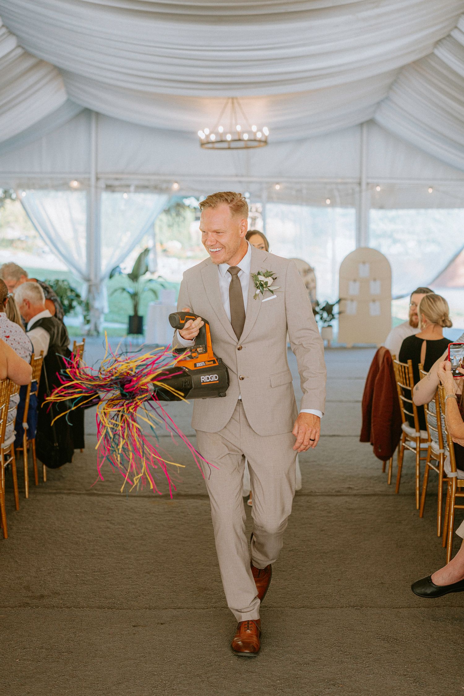 Person in gray suit walks down aisle in white tent reception venue with colorful streamers.