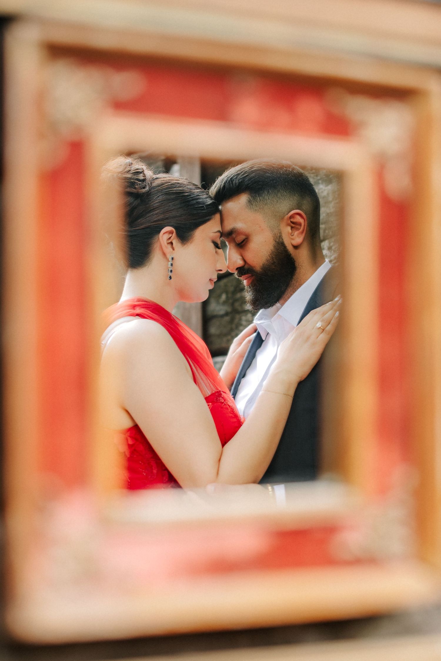 A romantic couple in red and blue outfits share an intimate moment framed through a window with orange tones.