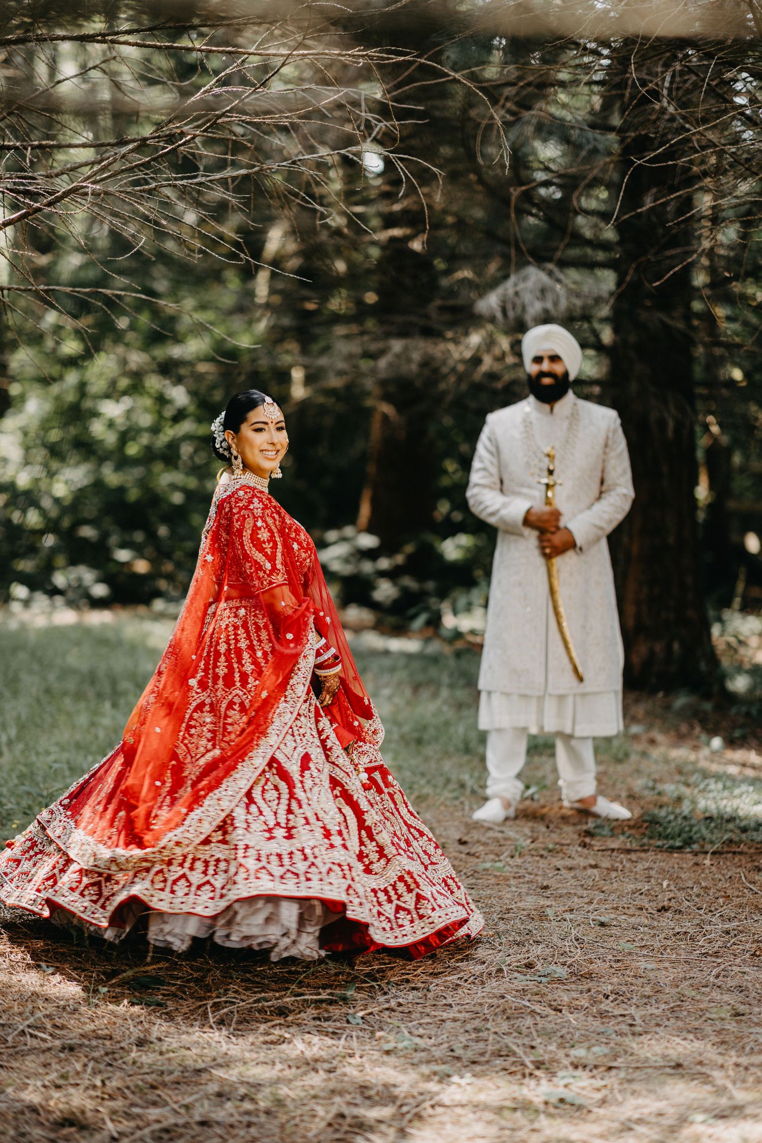 Traditional Indian wedding couple poses in outdoor setting with bride in red lehenga and groom in white sherwani.