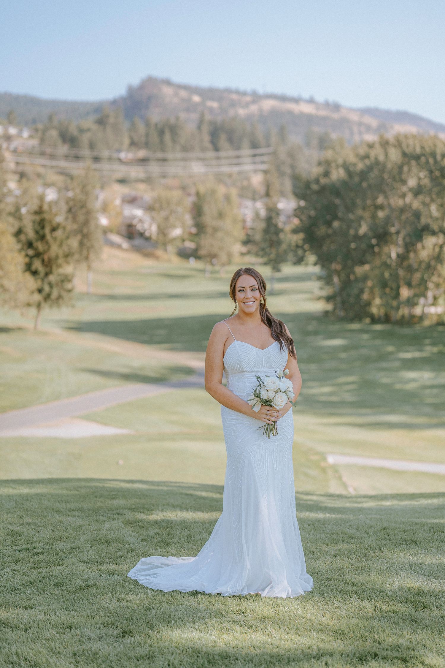 A bride in a white gown poses solo on a scenic golf course with mountains in the distance.