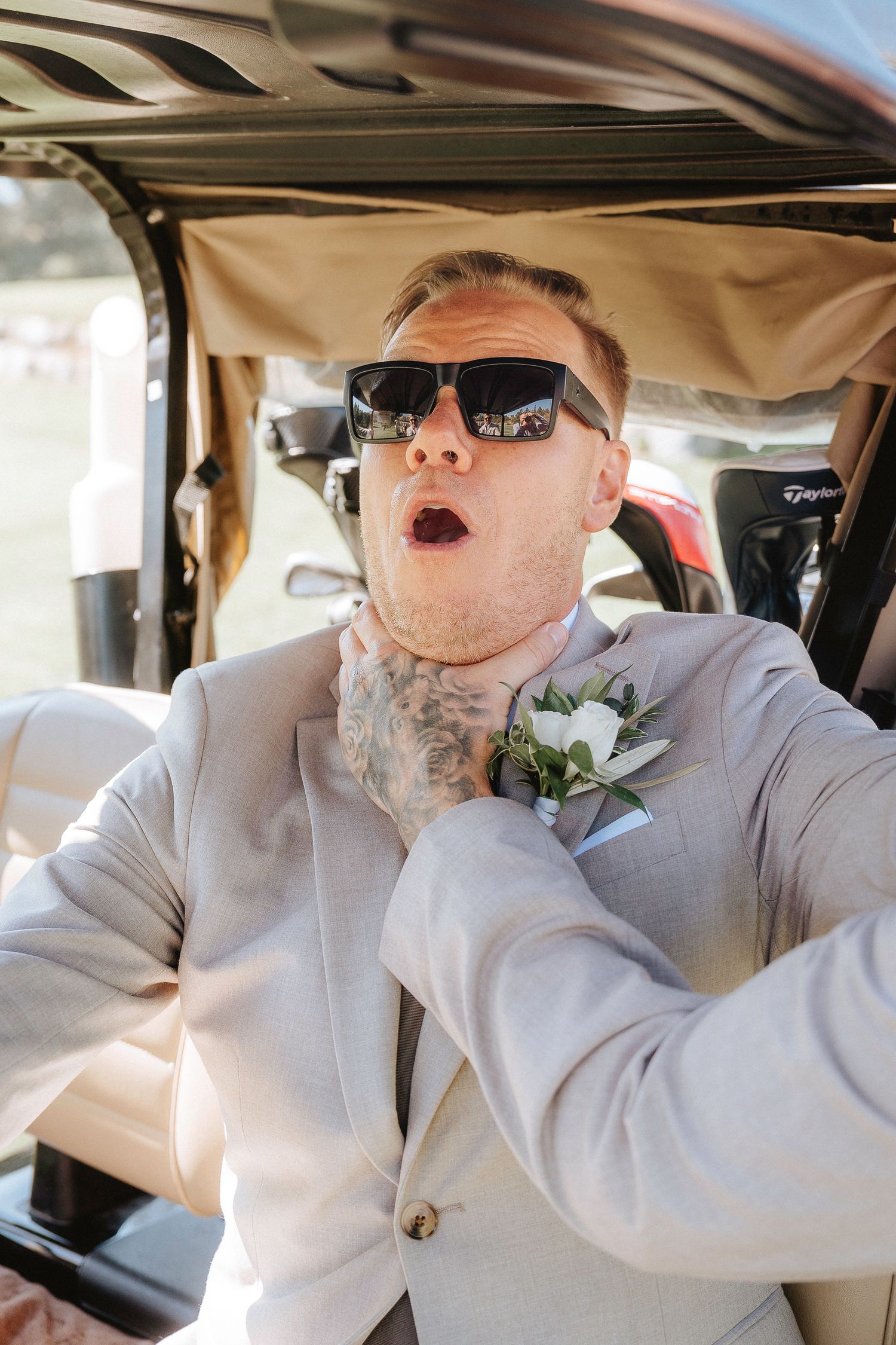 Wedding guest in light gray suit and sunglasses makes exaggerated expressions while seated in a car.