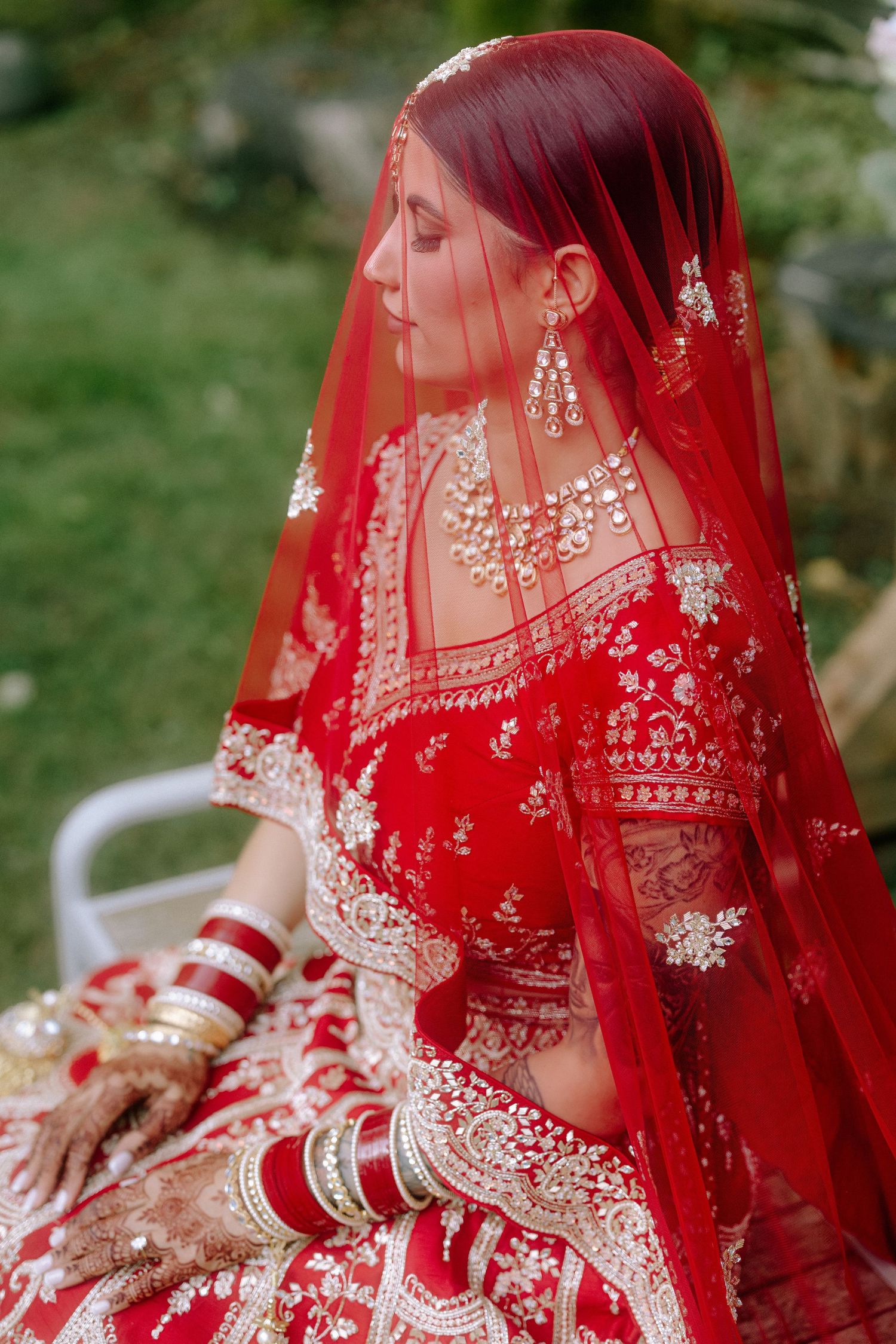 A traditional bride in a red wedding sari with intricate embroidery and veil sits displaying henna adorned hands.
