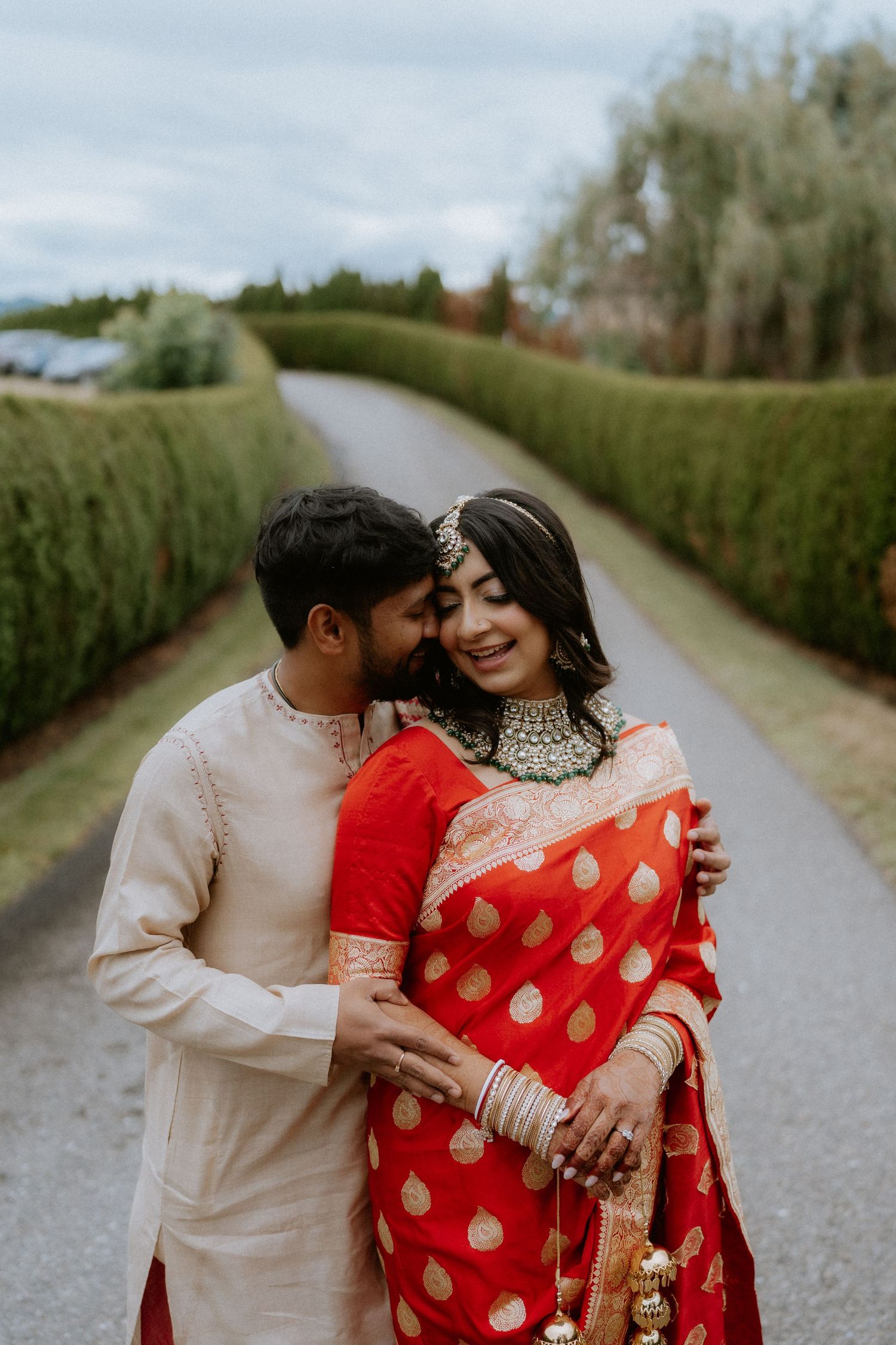 Romantic outdoor photoshoot featuring couple in traditional Indian wedding attire on a tree-lined path.